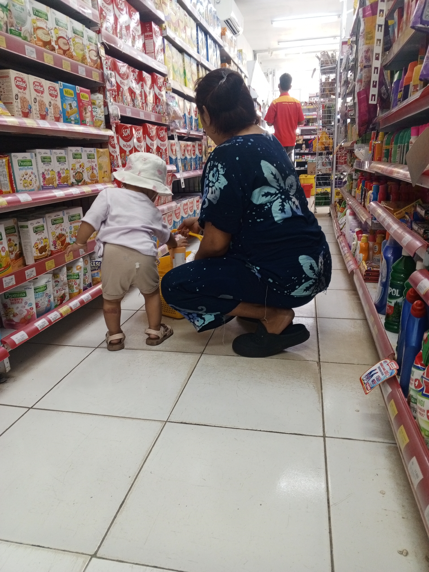This is a photograph taken indoors, likely in a convenience store or small supermarket located in Talun Kenas, Indonesia. The camera is positioned at a low angle, looking towards the back of a woman and a young child.
The woman is wearing a dark blue patterned dress and black slip-on shoes. She is crouched down, appearing to interact with the child, possibly helping them reach something or put something into a shopping basket, part of which is visible in her right hand.
The child is facing away from the camera, wearing a light-colored t-shirt, tan shorts, and sandals. They are standing and looking towards the store shelves. A light-colored bucket hat is on the child's head.
The store environment features rows of shelves filled with various products. On the left, there are shelves stocked with boxes of food items, seemingly cereal or formula, with prominent branding like "Promina" and "Milna". On the right, there are shelves displaying cleaning supplies and detergents in plastic bottles, with labels like "POL" and other unreadable text. The floor is tiled with a grid pattern. In the background, a man wearing a red and yellow shirt is visible, walking away from the camera, suggesting he is an employee. The lighting appears to be from overhead fluorescent fixtures.