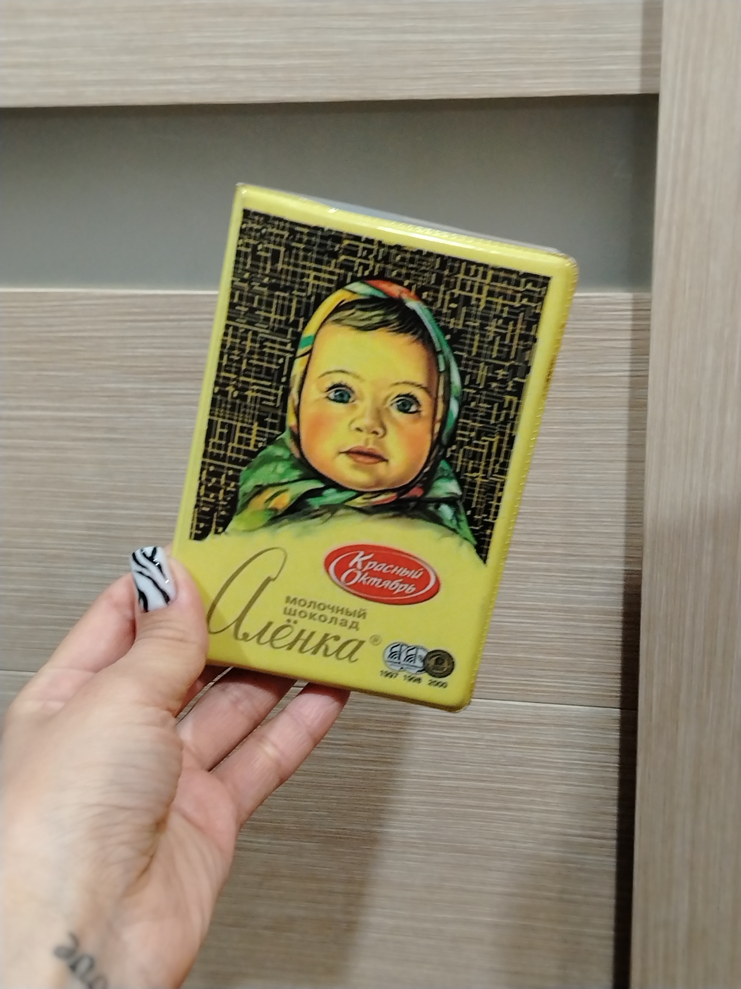 An indoor, eye-level shot shows a person's hand holding a rectangular bar of "Alenka" brand milk chocolate in Noviy, Russia. The chocolate bar packaging is primarily yellow and features the iconic illustration of a baby with light blue eyes wearing a colorful headscarf.
The visible text on the packaging includes "Алёнка" (Alenka) in large, stylized script, and "МОЛОЧНЫЙ ШОКОЛАД" (MILK CHOCOLATE) underneath it. The manufacturer's logo, "Красный Октябрь" (Krasny Oktyabr - Red October), is prominently displayed in a red oval. Below the logo, smaller text indicates years: "1997 1998 2000".
The hand holding the chocolate bar has manicured nails, with one visible thumb nail featuring a distinctive black and white zebra pattern. A partial tattoo, showing the numbers "200", is visible on the person's wrist. The background consists of a light-colored, horizontally-grained wooden or laminate surface, likely a door or cabinet, with a darker, recessed area behind the chocolate bar. The scene is well-lit, indicating an indoor setting.