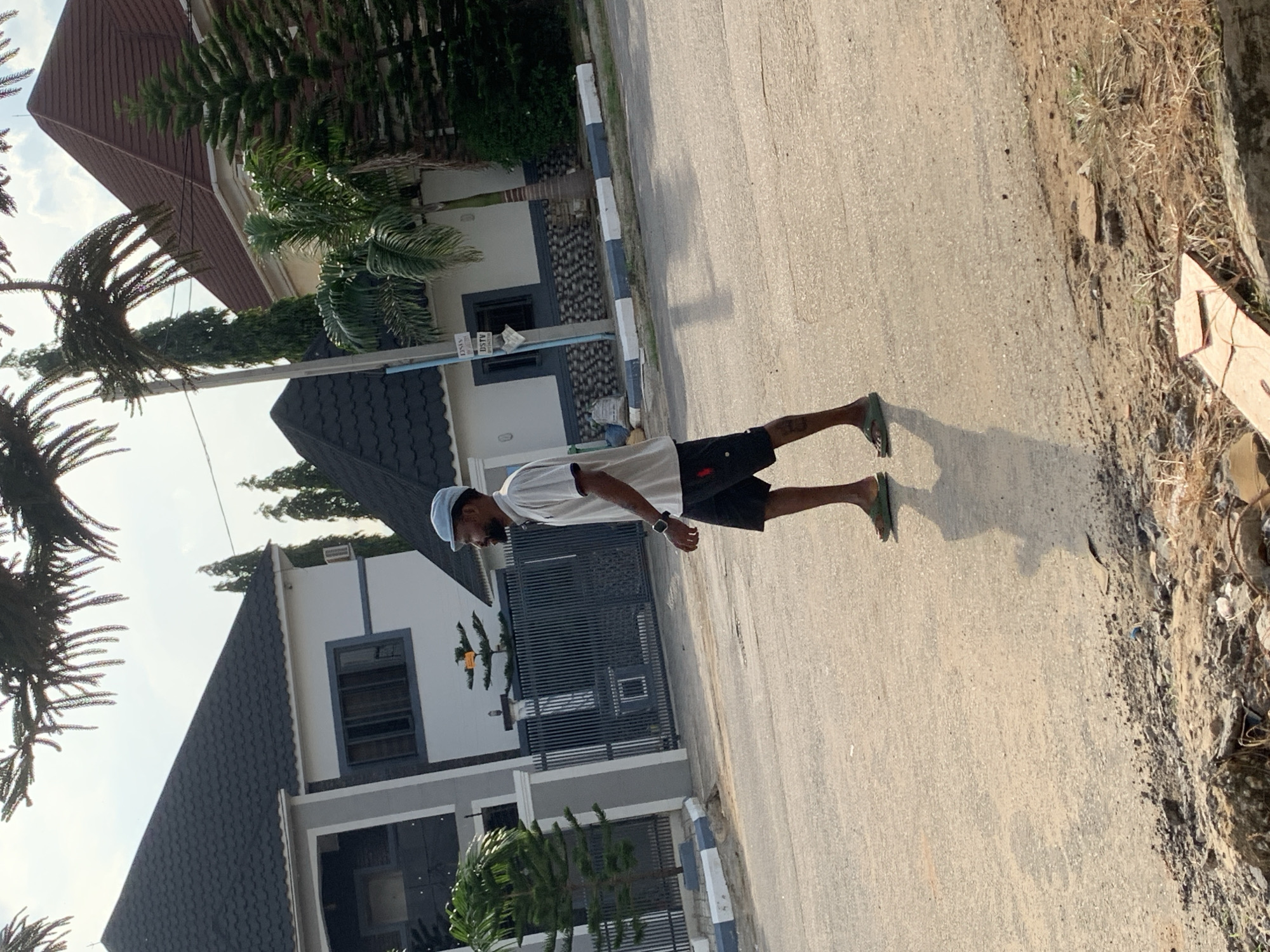 A medium-shot, full-body, side-profile photograph shows a man walking on a paved street in Abuja, Nigeria. The man is wearing a light blue cap, a white polo shirt, black shorts, and green flip-flops. He is walking from left to right, with his head turned to the right and his arms at his sides. His shadow is cast on the street to his left.
In the background, on the left side of the image, is a modern, two-story house with a dark gray roof, white walls, and dark gray window frames. A dark gray metal fence encloses the property. A small green tree stands in front of the fence.
To the right of the first house, there is another building with a dark gray, pitched roof and white walls with a dark gray stone-like base. A palm tree is visible in front of this building. Further to the right, another house with a reddish-brown, pitched roof and white walls can be seen, with pine trees in front of it.
The street is paved with asphalt and shows signs of wear. The area to the left of the street is unpaved, with patches of dry grass, dirt, and debris, including what appears to be broken concrete or rocks. The sky is visible at the top of the image, appearing partly cloudy. The overall lighting suggests it is daytime, likely late afternoon given the long shadows.