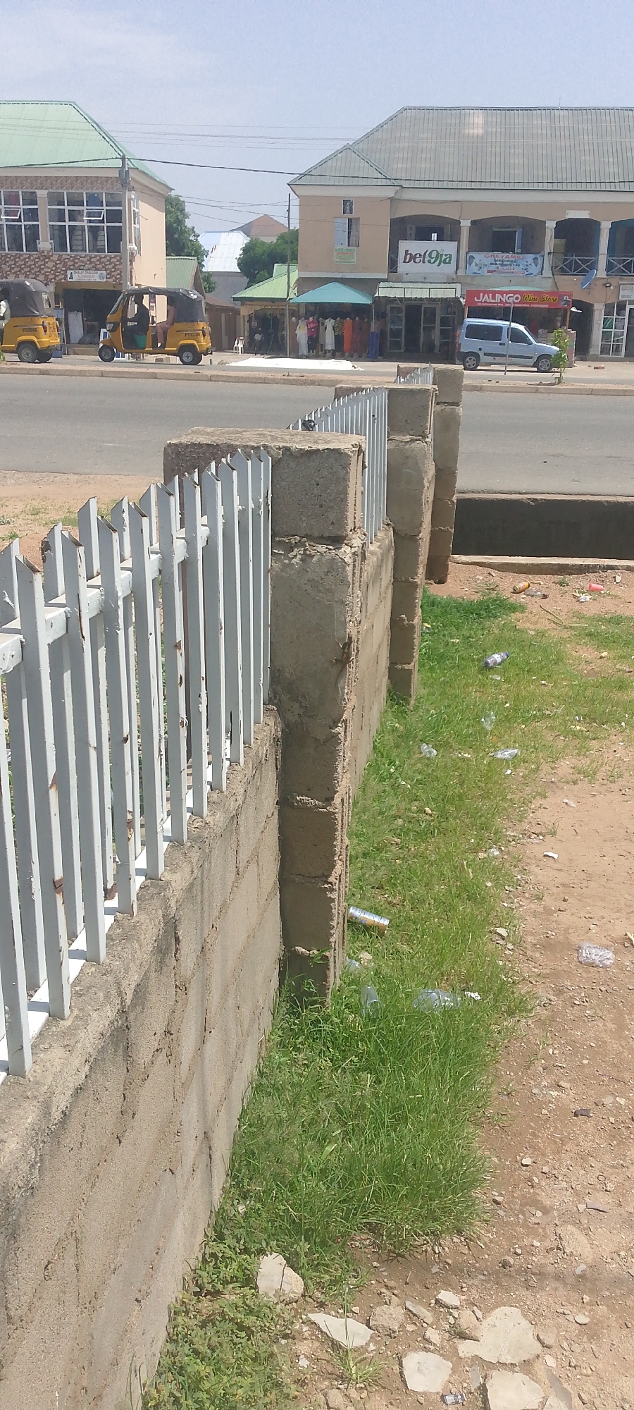 This image captures a street scene in Jalingo, Nigeria, likely during the daytime under clear skies. The foreground features a low, weathered brick wall topped with a white metal fence, partially obscuring the view. Beyond the fence, a paved road runs horizontally across the frame, with a dark, rectangular drainage channel running alongside the road.
On the other side of the road, a row of buildings lines the street. To the left, a building with a light-colored facade and a green roof features several windows on its upper level and what appears to be shops on the ground floor. In front of these shops, two yellow auto-rickshaws, known as "keke napep," are parked or moving. One has a driver visible.
Further down the street, another building with a similar architectural style is present. This building has a larger facade, also light-colored, with an arched upper story. Various signs are visible on this building. One prominent sign reads "bet9ja." Another sign beneath it says "JALINGO Wine Store." Below these, a smaller sign is partially obscured. In front of this building, awnings extend over shop entrances, and mannequins display colorful clothing. Several people are seen in front of these shops, suggesting commercial activity. A light blue van is parked near the "JALINGO Wine Store."
The ground along the roadside, particularly to the right of the wall and fence, is covered in patchy green grass and scattered debris, including plastic bottles and cans, indicating a slightly unkempt area. The overall impression is a bustling urban environment with commercial activity and transportation common in many Nigerian towns.
