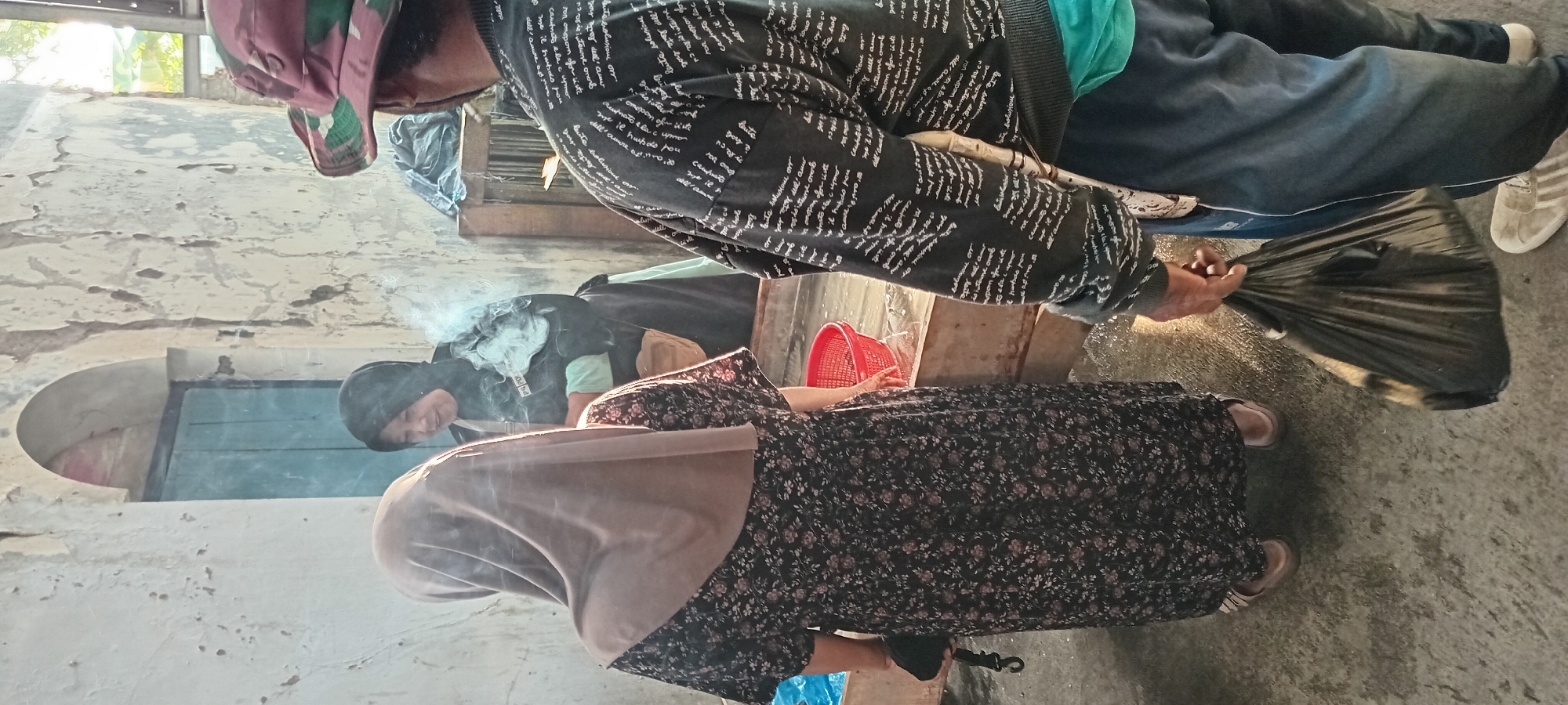 In a semi-outdoor setting, possibly a market or a stall in Sindangwangi, Indonesia, three individuals are present. The focus is on a woman in the foreground wearing a brown hijab and a floral print dress. She is facing away from the camera, looking towards a second woman. This second woman, wearing a black hijab and a green garment, is partially visible, smiling and in the process of smoking. White smoke billows from her mouth, obscuring part of her face. A third person, a man wearing a camouflage-patterned hat and a dark patterned sweater, stands to the right, facing the two women. He is holding a black plastic bag in his right hand. The environment appears rustic, with a rough, textured wall behind the individuals and a blue wooden door in an arched opening. A red plastic basket is visible on a wooden counter or surface, along with some water. The ground is concrete and wet, reflecting light. The scene suggests a casual, possibly everyday interaction.