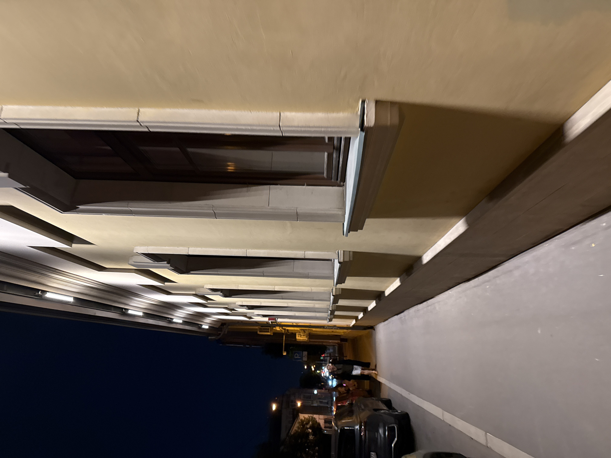 The image presents a detailed nighttime street scene in Moscow, Russia, captured from a low, oblique perspective looking down a street and an illuminated building facade.
On the right, a multi-story building with a pale yellow or cream-colored stucco facade dominates the foreground and extends into the middle distance. Below its prominent cornice, a series of linear, horizontally mounted LED light fixtures uniformly illuminate the facade with a cool white light. The building features numerous recessed rectangular windows with dark brown or black frames, each bordered by light-colored decorative stone or stucco surrounds and substantial sills. Reflections from indoor lighting are visible within the windowpanes.
A smooth, grey concrete sidewalk runs parallel to the building, extending from the bottom center into the distance. A distinct white line is painted along the edge of the sidewalk, marking a clear division between the pedestrian path and the street parking area.
To the left of the sidewalk, a dark grey or black SUV is parked, with its front bumper and part of its wheel well visible in the lower left corner. The vehicle's license plate is partially visible, displaying the "777" region code, which corresponds to Moscow registration. Further down the street, the rear section of a red vehicle is also parked.
One individual is observed walking on the sidewalk in the middle distance, moving away from the camera. They are wearing a dark upper garment, possibly a jacket, and light-colored shorts or a skirt. Their facial features are indistinct due to the distance and ambient lighting. In the background, several streetlights emit a warm, yellow-orange glow, and a blue "P" parking sign is visible. The sky overhead is a deep, dark blue, characteristic of a clear night.