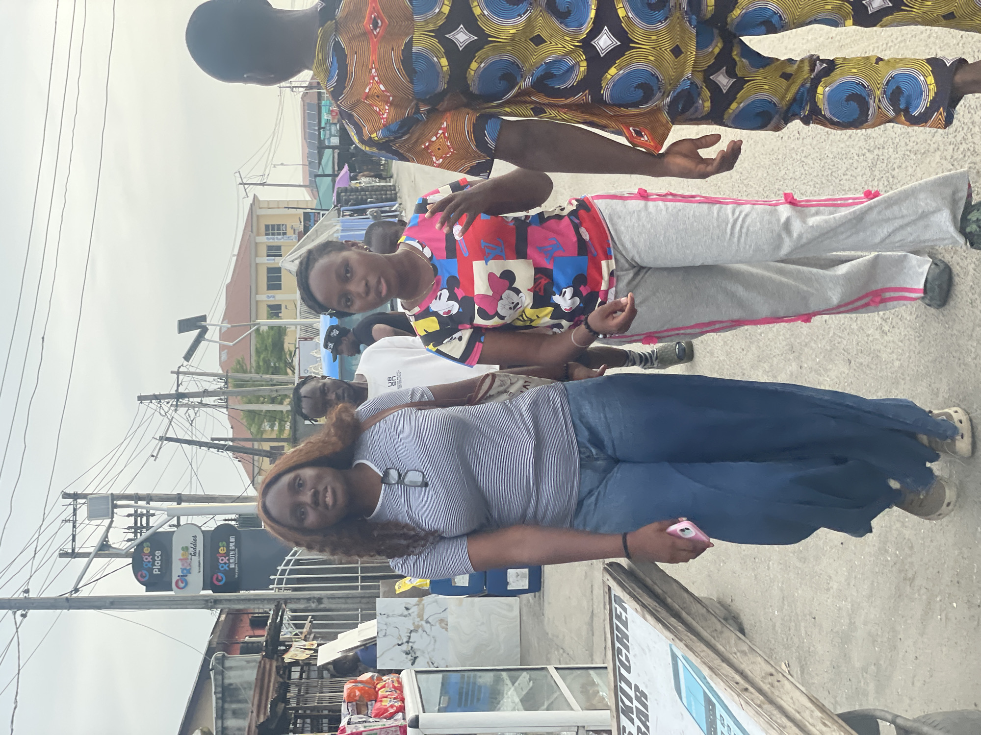 This outdoor street scene, set in an urban environment likely in Ado, Nigeria, features several individuals amidst a backdrop of buildings, utility poles, and commercial signs. The sky, partially overcast, suggests it is daytime.
In the foreground, on the left, a woman with long, wavy brown hair and glasses resting on her striped short-sleeve shirt stands, wearing blue pants and beige slip-on shoes, holding a pink phone. Next to her, in the middle of the frame, is a young person sporting a brightly colored t-shirt adorned with cartoon characters (Mickey and Minnie Mouse) and motifs resembling Louis Vuitton branding, paired with light grey sweatpants featuring pink side stripes. Their hands are slightly raised. To the far right, with their back partially turned, another person is visible, wearing a vibrant, intricately patterned shirt characteristic of West African Ankara fabric. Further in the background, additional people are partially obscured.
The setting appears to be a busy commercial area. Prominently displayed are three stacked signs for "Giggles Place," "Giggles Facilities," and "Giggles BEAUTY SALON," each bearing the tagline "your go-to hub." Below these, numerous utility poles crisscrossed with wires are visible. In the background, buildings with yellow walls and brown roofs line the street. In the lower left foreground, a display with white and grey marble-patterned tiles is present, alongside a partial sign advertising "KITCHEN BAR." Various items, possibly merchandise or groceries in bags, are also visible on stands near the ground. The individuals appear to be engaged in typical street activity, such as walking or stopping to observe their surroundings.