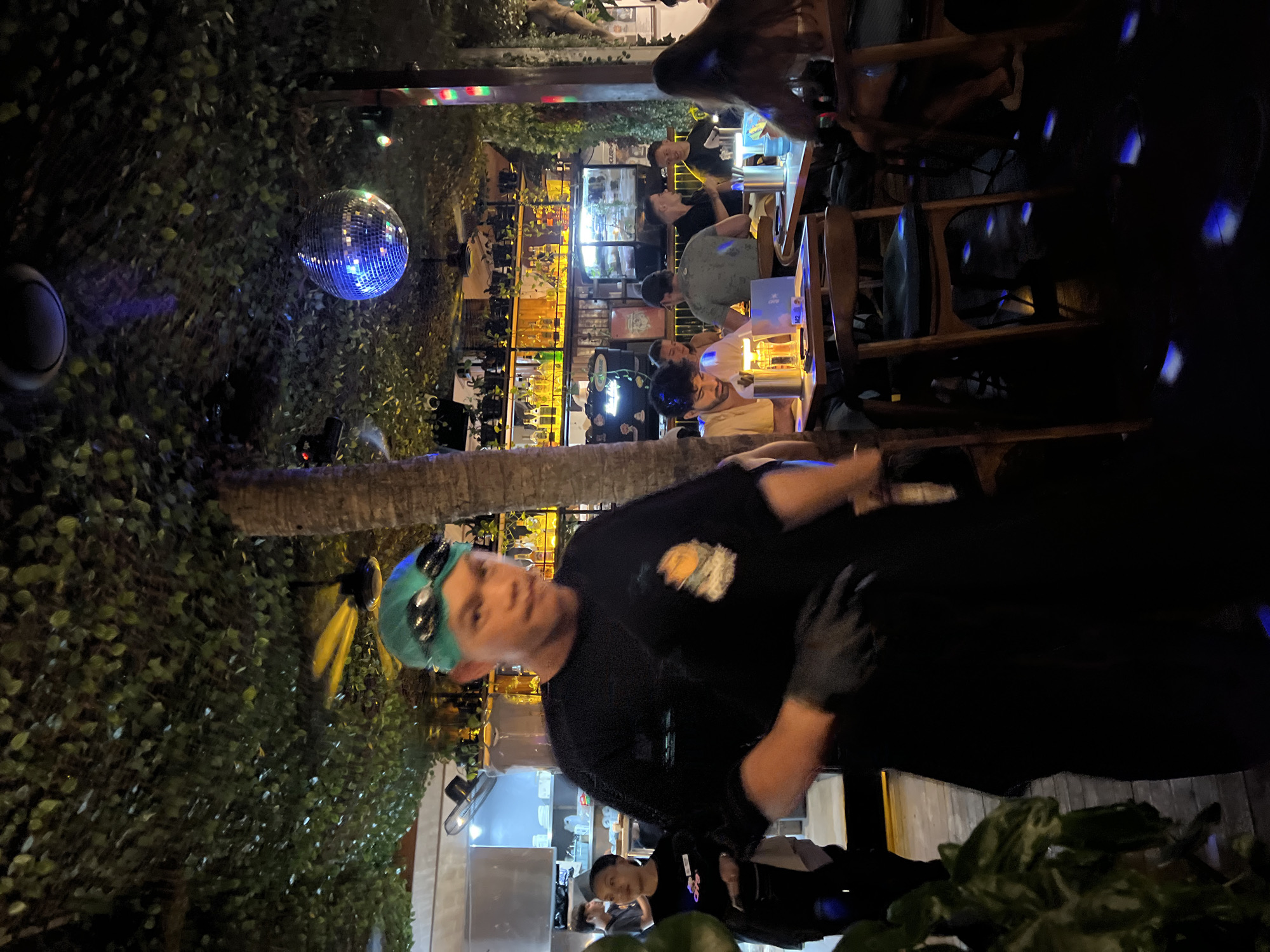 The image depicts an indoor scene, likely a bar or restaurant, with a disco ball hanging from the ceiling and illuminated by ambient lighting. A person wearing a teal swimming cap and goggles is the main subject in the foreground, standing and facing the camera with a slightly blurred expression. They are wearing a black t-shirt and black gloves. Behind this individual, a woman in a black t-shirt is visible, and further back, several other patrons are seated at tables, some engaged in conversation and dining. The environment is decorated with abundant greenery, creating a lush and somewhat enclosed atmosphere. The presence of a disco ball suggests a lively, possibly entertainment-focused venue. The specific location within Mengwi, Indonesia, cannot be definitively determined from the image alone, as there are no explicit geographic markers. However, the overall ambiance and decor might be characteristic of establishments in tourist areas of Bali, Indonesia.