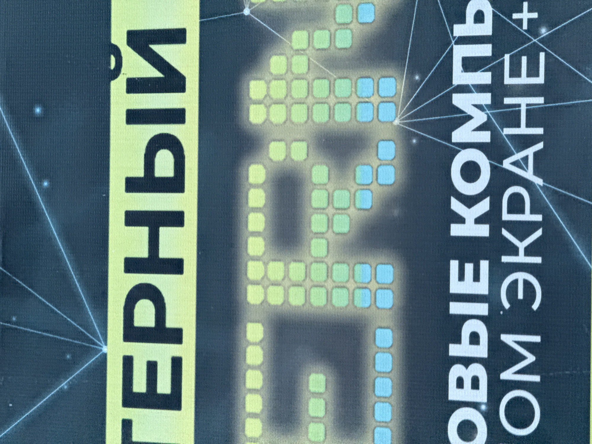 The image shows a close-up of a promotional poster or sign, likely related to computer technology or gaming. The visible text is in Russian. On the left, the word "ГЕРНЫЙ" ("GERNYY") is printed vertically in black letters on a yellow background. To the right, a pixelated graphic or logo consisting of green and blue squares forms a word or symbol. Further to the right, white text reads "ОВЫЕ КОМПЬ / ОМ ЭКРАНЕ+". The background is a dark blue with white lines and dots that may represent a digital network or schematic. The text and imagery suggest an advertisement for computer technology or gaming. The city, Votkinsk, Russia is not discernible from the image itself.