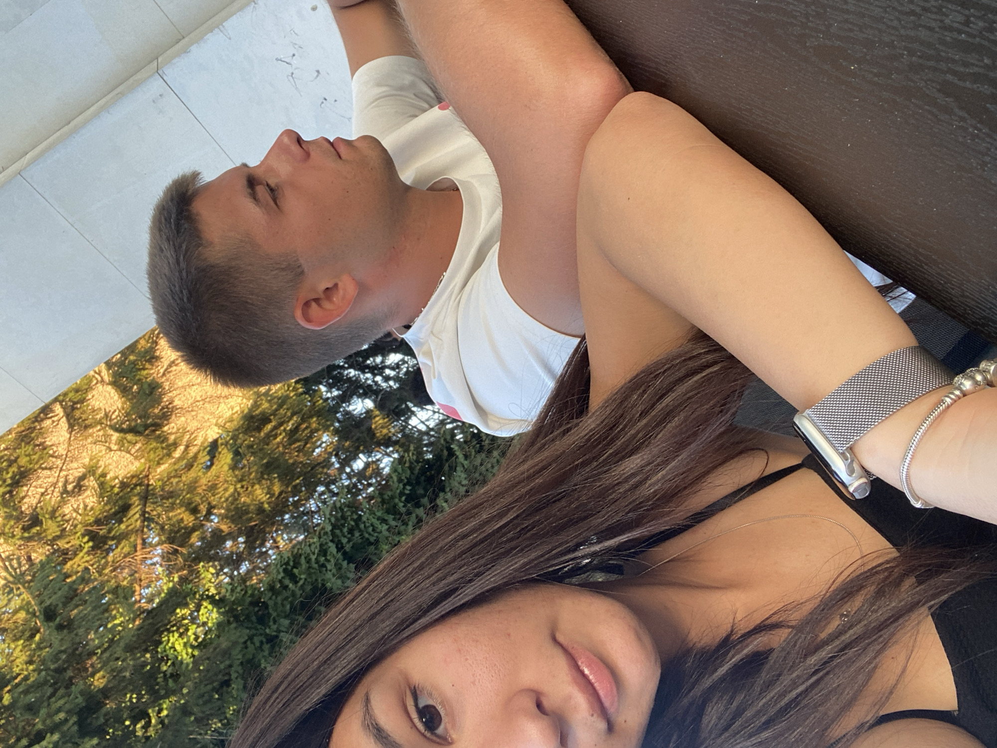 A close-up, candid selfie shows a young woman in the foreground and a young man in the background. The woman, positioned on the left, has long, dark brown hair that cascades over her shoulders. Her right arm is bent, resting on a dark brown surface, and she wears a silver-banded smartwatch and a beaded bracelet. She is looking towards the camera with a slight smile. The man, to the right and slightly behind her, has short, light brown hair and is wearing a white t-shirt. He is looking off to the right, with his arms resting on the same dark brown surface. They are seated outdoors, with lush green trees and sunlight visible in the background, suggesting a park or garden setting. The overall lighting indicates it is daytime, likely late afternoon due to the warm tones of the sunlight. The location is identified as Sevlievo, Bulgaria.