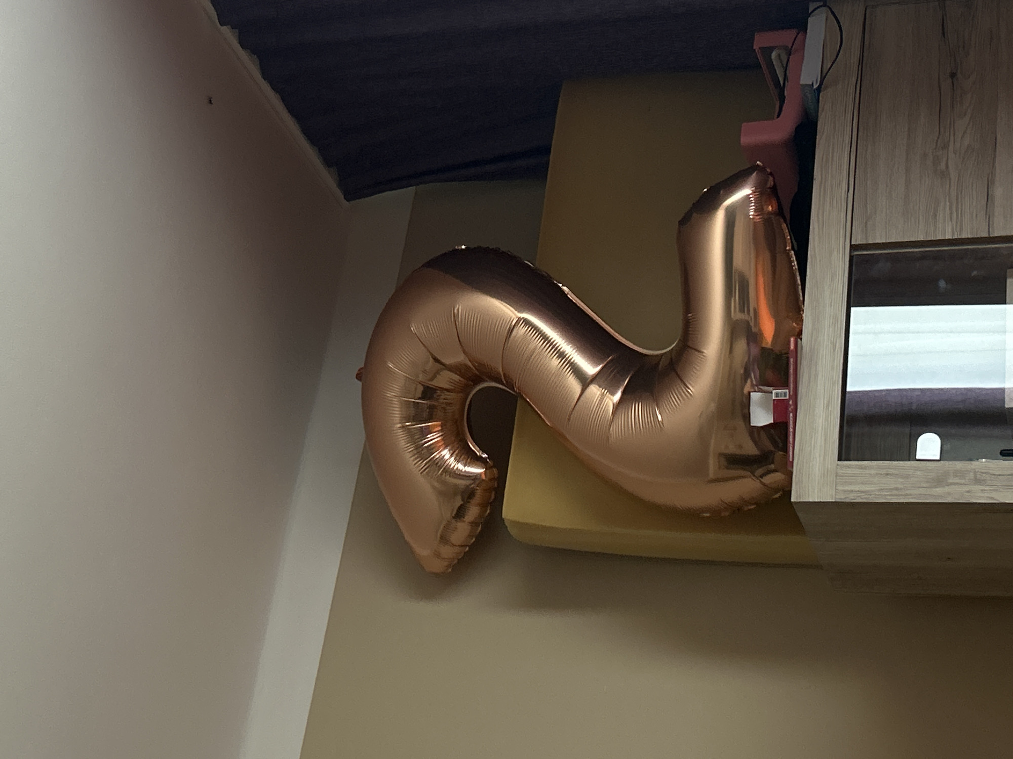 This media file captures a close-up view of a rose gold number "2" balloon, inflated and casting a slight shadow. The balloon is the primary subject, positioned in the foreground and dominating the frame. It rests on what appears to be a yellow cushioned surface, possibly a sofa or chair.
In the background, a light-colored wall forms the left boundary of the image, transitioning to a darker, angled surface at the top, which could be a ceiling or an overhang. To the right, a wooden cabinet with a glass-fronted compartment is visible. Within the cabinet, a white, arch-shaped light is illuminated. The overall lighting suggests an indoor setting, likely during daylight hours, though artificial light from the cabinet is also present.
A pink object, possibly another decorative item or part of a toy, is partially visible behind the balloon and next to the cabinet. There is a small piece of white paper attached to the balloon with some red text and a barcode, though the text is not legible. No people are visible in the image. The scene is static, with no discernable activity taking place. The composition, with the balloon prominently featured and the background elements providing context, suggests a celebratory or decorative arrangement. The location is Teplice, Czechia.