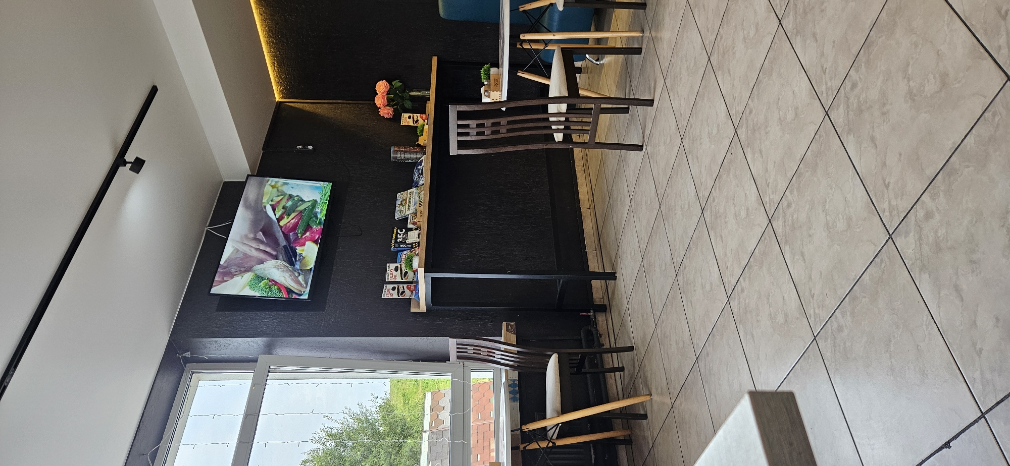The image depicts the interior of a cafe or restaurant. A flat-screen television is mounted on a dark-colored wall, displaying an image of food preparation. Below the television, a wooden shelf holds various items, including books and decorative elements. Several chairs are positioned around tables. A large window on the left side of the frame looks out onto an outdoor scene with trees and buildings. The floor is tiled with a light beige pattern. The overall impression is of a modern and casual dining establishment. The specific location in Vitsebsk, Belarus cannot be determined from the image alone.