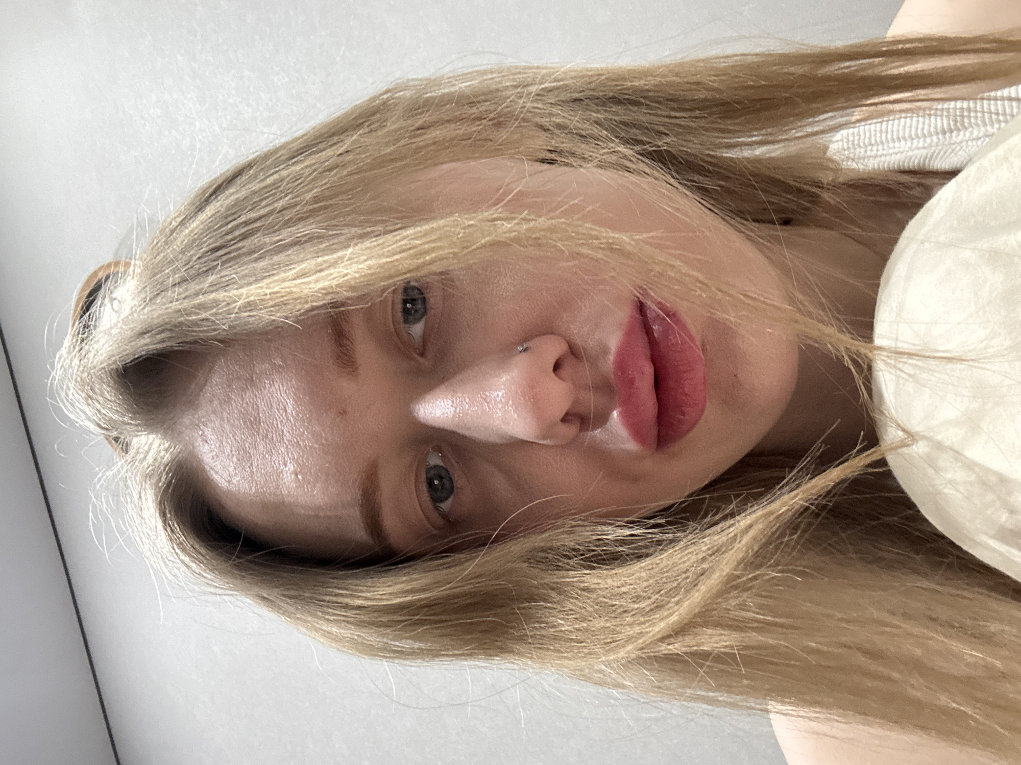 A close-up, head-and-shoulder portrait depicts a light-skinned female individual within an indoor setting in Rostov-on-Don, Russia. The subject has long, straight blonde hair, with some strands obscuring parts of her face. Her eyes are blue-grey and directed towards the viewer. Her lips are full with a natural or applied pinkish-red color. A small metallic stud is visible in her left nostril. The skin on her forehead and around her nose appears somewhat reflective. A light-colored garment, possibly a strap or tank top, is visible on her right shoulder, and another light-colored, possibly fabric object, is present at the bottom center of the frame. The background is a plain, light grey wall with a thin dark horizontal line at the top.
