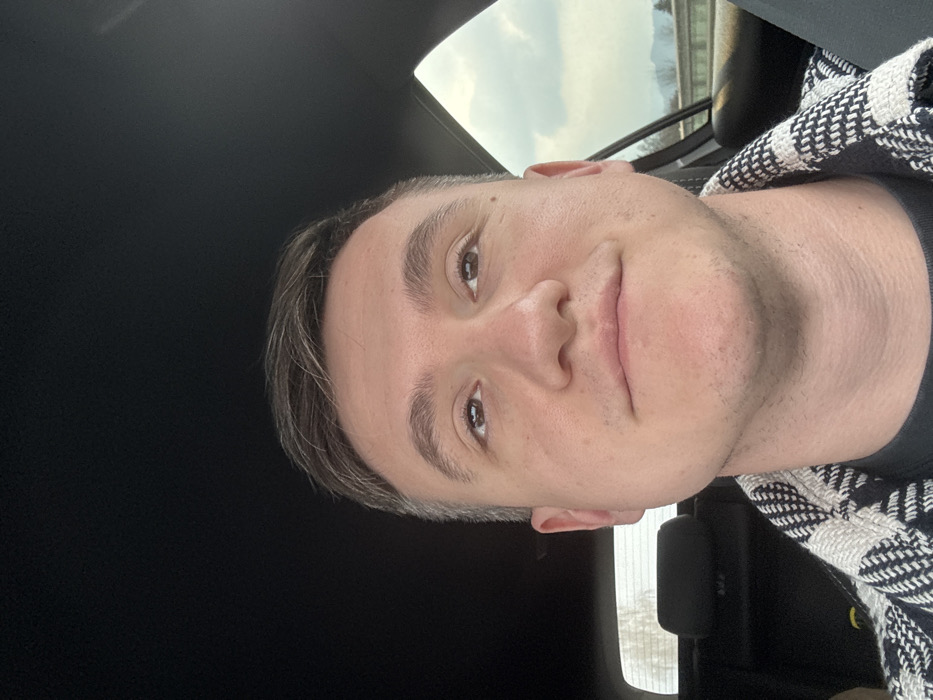 The image captures a close-up, selfie-style view of a male individual seated within a vehicle. The subject is a light-skinned adult with short brown hair, wearing a black and white patterned outer garment over a dark undershirt. His eyes are directed upwards and slightly to his left, with a neutral to slight smiling expression. The vehicle's dark headliner is visible above him. Through the window to his right, a cloudy daytime sky is visible above distant hills or mountains, with a grey barrier or guardrail present below. To his left, a black headrest is visible in the background, partially obscuring the view through a rear window. No specific landmarks are present in the image to confirm the location as Aichdorf, Austria.