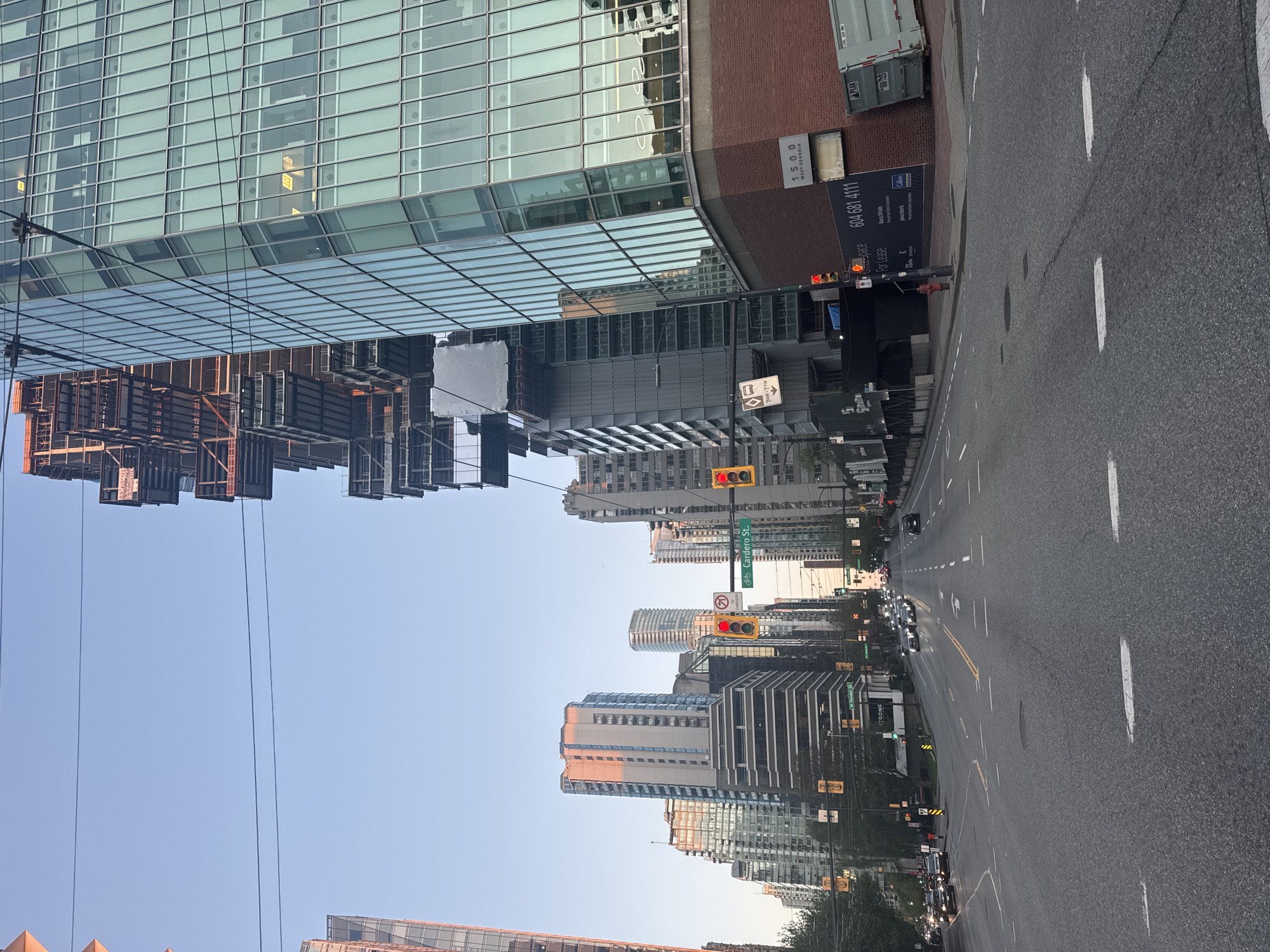 This image presents a daytime urban street scene in Vancouver, Canada, looking down a wide multi-lane road flanked by modern high-rise buildings under a clear blue sky. The perspective extends into the distance, revealing a continuous stretch of city infrastructure.
On the left side of the road, a prominent skyscraper with a reflective glass facade dominates the foreground, mirroring adjacent structures and the sky. Next to it, a building featuring a brick upper section displays the address "1500 WEST GEORGIA" and an advertisement for "Office space For Lease" with the contact number "604 681 4111." The lease sign also mentions "Colliers International" and agents "Mara Paplo" and "James Norris." A white shipping container labeled "PEAK" and "690-7225" is visible near this building.
The road itself is paved with asphalt, marked by clear white dashed lines indicating lanes and solid yellow lines for separation. Multiple vehicles are visible further down the street, suggesting active traffic. Several traffic lights are present, with at least two showing red, indicating a stop or caution for vehicles. A green street sign clearly identifies "Cardero St." An additional sign with a parking symbol indicates a time restriction "7AM-7PM" with an arrow pointing right, likely signifying a no-parking or tow-away zone. Overhead, power lines crisscross the sky. Other buildings with diverse architectural styles, including modern glass and concrete designs, line both sides of the street as it recedes into the background. The word "STRONG" is also visible on a sign in the distance, possibly a business name.