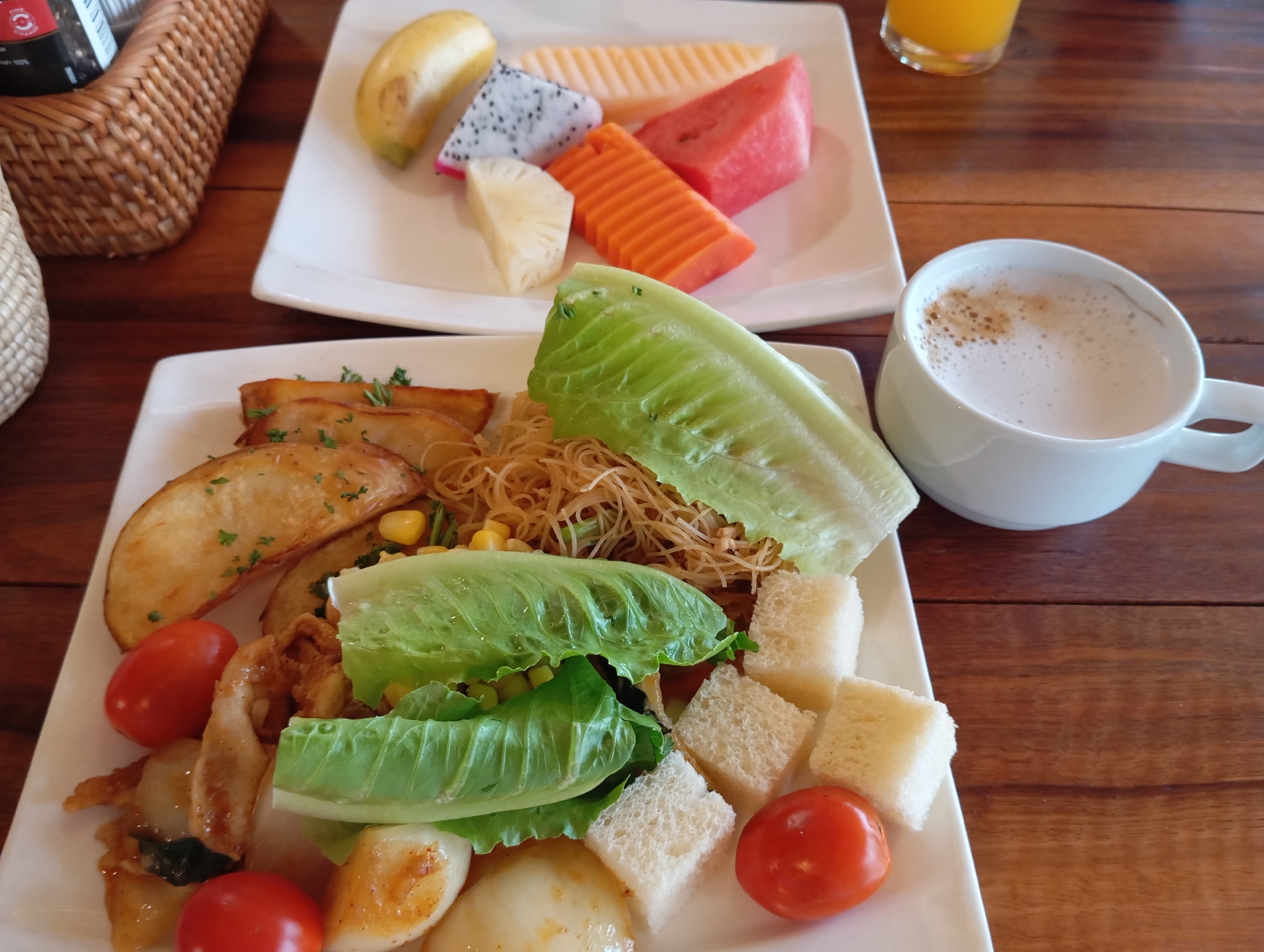 In Karon, Thailand, a table laden with breakfast items is captured from a top-down perspective. The focus is on two white rectangular plates of food set on a polished wooden surface. The plate in the foreground contains a diverse spread: roasted potato wedges sprinkled with herbs, a pile of thin noodles, corn kernels, two large pieces of lettuce, cubed white bread, cherry tomatoes, and sliced hard-boiled eggs. A white mug filled with a foamy coffee beverage sits to the right of this plate. The background plate showcases a colorful assortment of sliced fruits: a banana, dragon fruit, melon, papaya, and pineapple. A partially visible glass of orange juice is also present in the background, along with a woven basket. The lighting suggests an indoor setting, possibly a restaurant or hotel dining area.