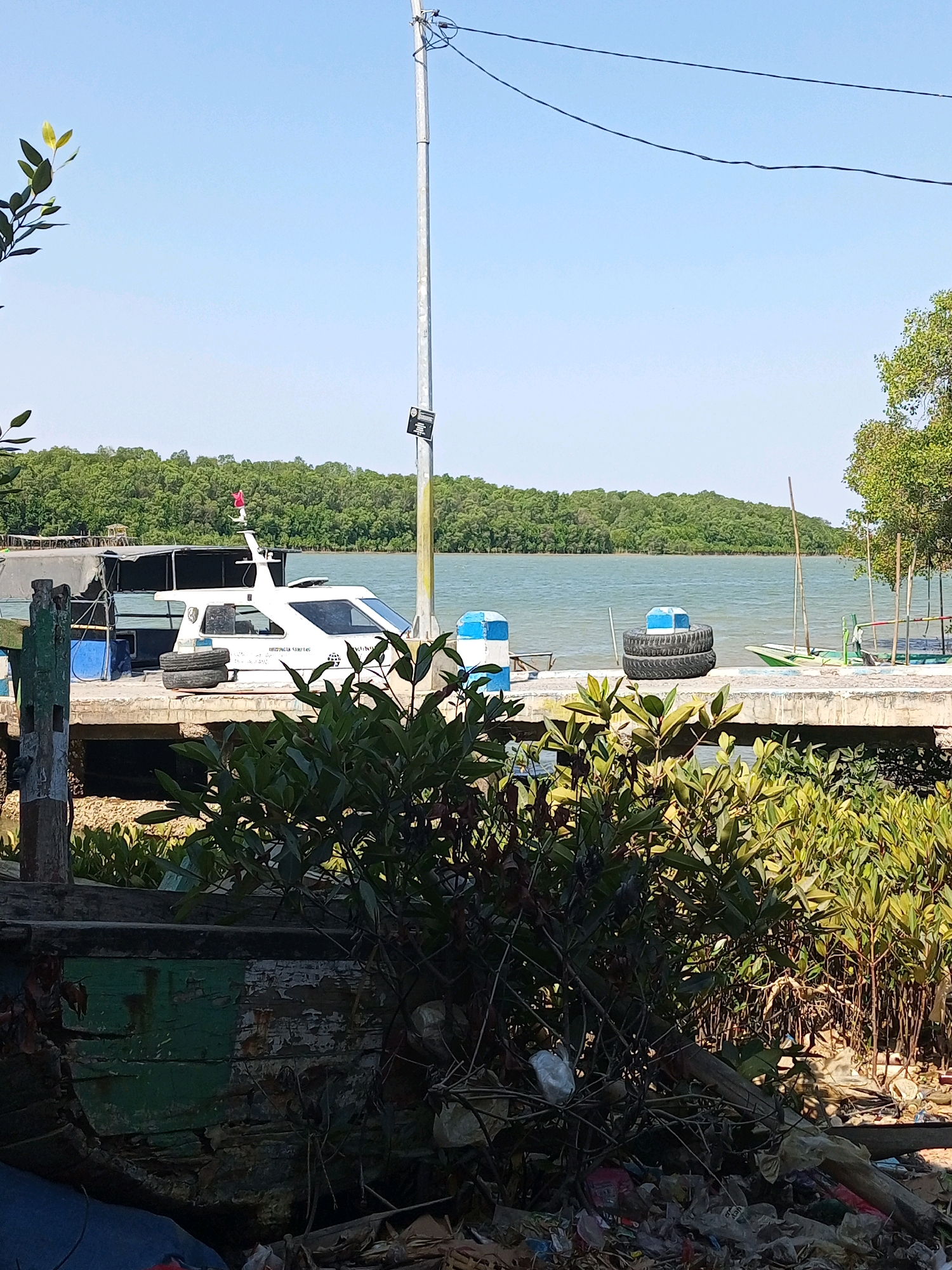 The image depicts a waterfront scene on a sunny day. In the foreground, there is a pile of garbage and overgrown green plants. Behind the plants, a weathered wooden structure is visible, painted green and white, with peeling paint. A concrete pier extends into the water, where a white boat with a dark interior and tinted windows is docked. Two tires are stacked on the pier next to the boat. Further out on the water, a lush green treeline forms the horizon. A tall metal pole with electrical wires runs vertically through the scene, extending upwards into a clear blue sky. A small red flag is mounted on the boat. To the right, another pier juts out into the water, with a green boat and some wooden poles. The water is calm and appears slightly choppy.