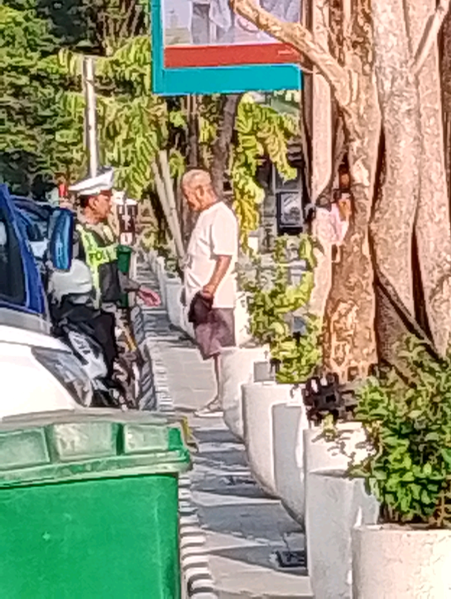 This image captures a street scene in Probolinggo, Indonesia, likely on a sunny day. In the foreground, a green trash bin with a reflective top is visible on the left. To the right of the trash bin, a police officer in uniform, including a white hat and a reflective vest with "POL" visible, is seated on a motorcycle. The officer is interacting with an older man, who is standing and holding a dark cloth in his right hand. The man is wearing a light-colored t-shirt and shorts. The background features a large tree with textured bark and lush green foliage. Several large, white planters with greenery are arranged along the sidewalk. The overall setting appears to be an urban street with elements of landscaping.