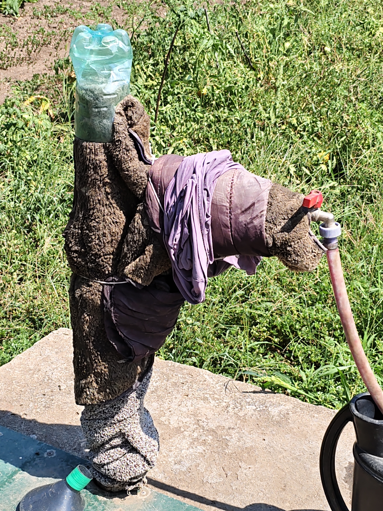 The image depicts a makeshift water dispenser in Nizhnedonskoy, Russia. A repurposed plastic bottle filled with water is positioned on top of a rough, textured post, which is wrapped in dark fabric and secured with white cord. A purple cloth is draped over the fabric. A garden faucet is attached to the side of the post, with a pink hose leading from it into a black watering can. The post stands on a concrete base, with green grass and foliage visible in the background. A black plastic bottle with a green cap lies on its side near the base of the post.