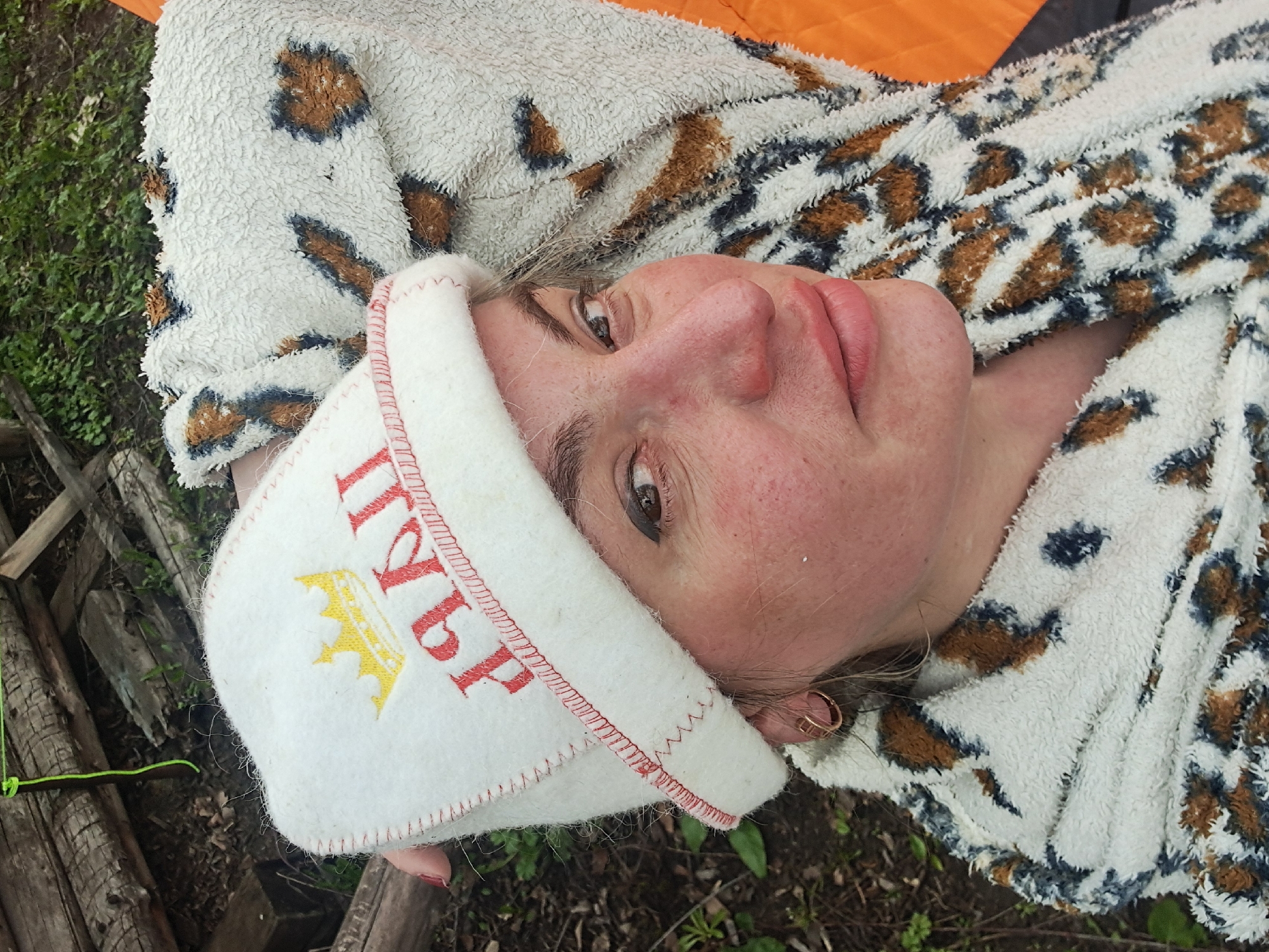 A middle-aged woman is seen lying down outdoors in Полянки, Russia. She is wearing a white terrycloth sauna hat with a red inscription, partially obscured, but featuring a yellow crown. The hat's trim is stitched in red. She is wrapped in a white and brown patterned fleece blanket. Her head is turned towards the camera. The background includes greenery, possibly grass and foliage, and fragments of what appear to be rough-hewn wooden structures or logs. Part of an orange material is visible in the upper portion of the image.