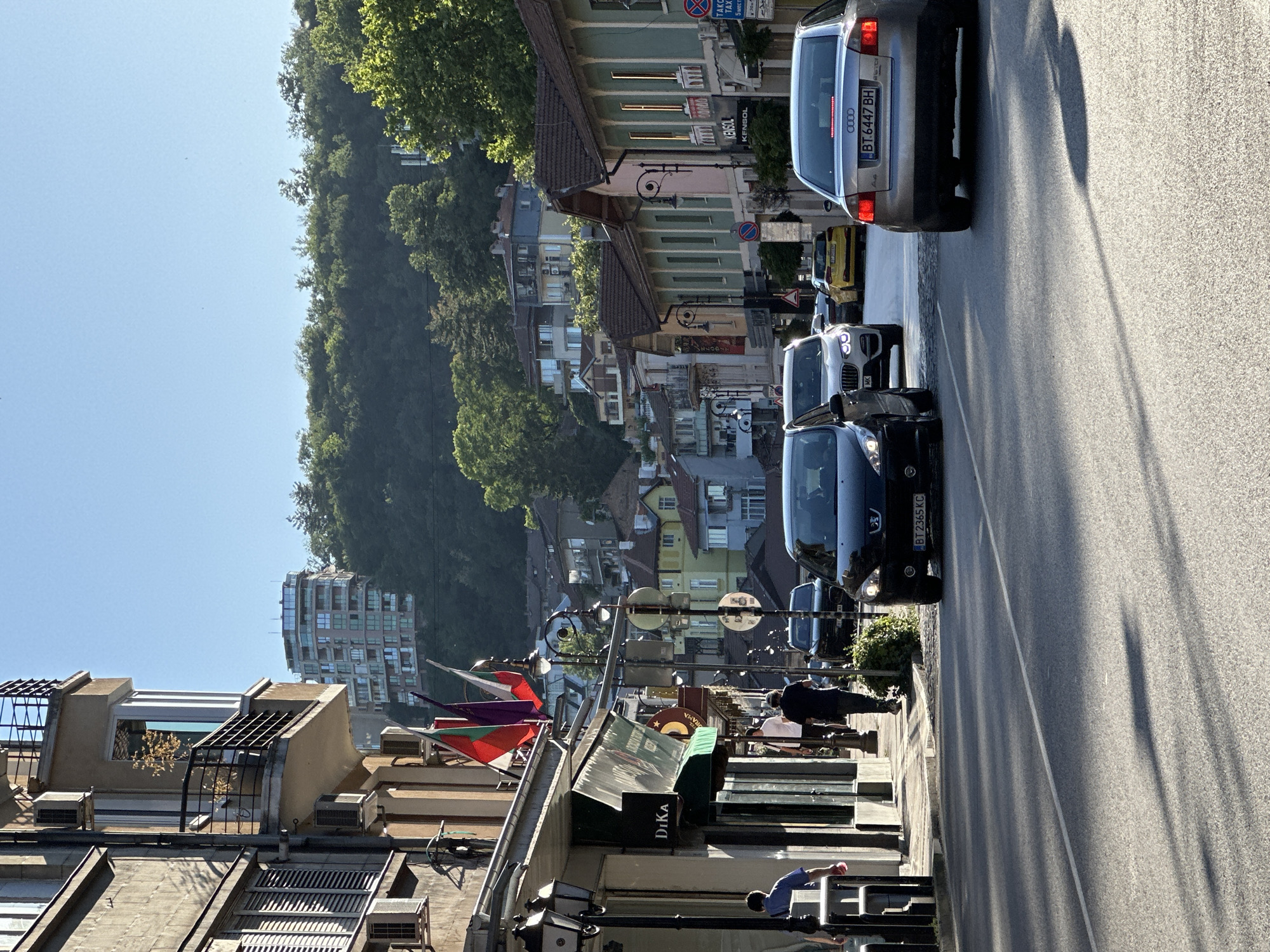The image depicts a street scene in Veliko Tarnovo, Bulgaria, on a sunny day. The street is lined with buildings of various architectural styles, some with balconies and awnings. Several cars are visible on the road, including a silver Audi, a black Peugeot, and a white BMW SUV. People are walking on the sidewalk, some entering and exiting shops. Bulgarian flags are displayed on poles in front of one of the buildings. In the background, the lush green hills of Veliko Tarnovo rise, dotted with more buildings and trees. The sky is clear and blue.