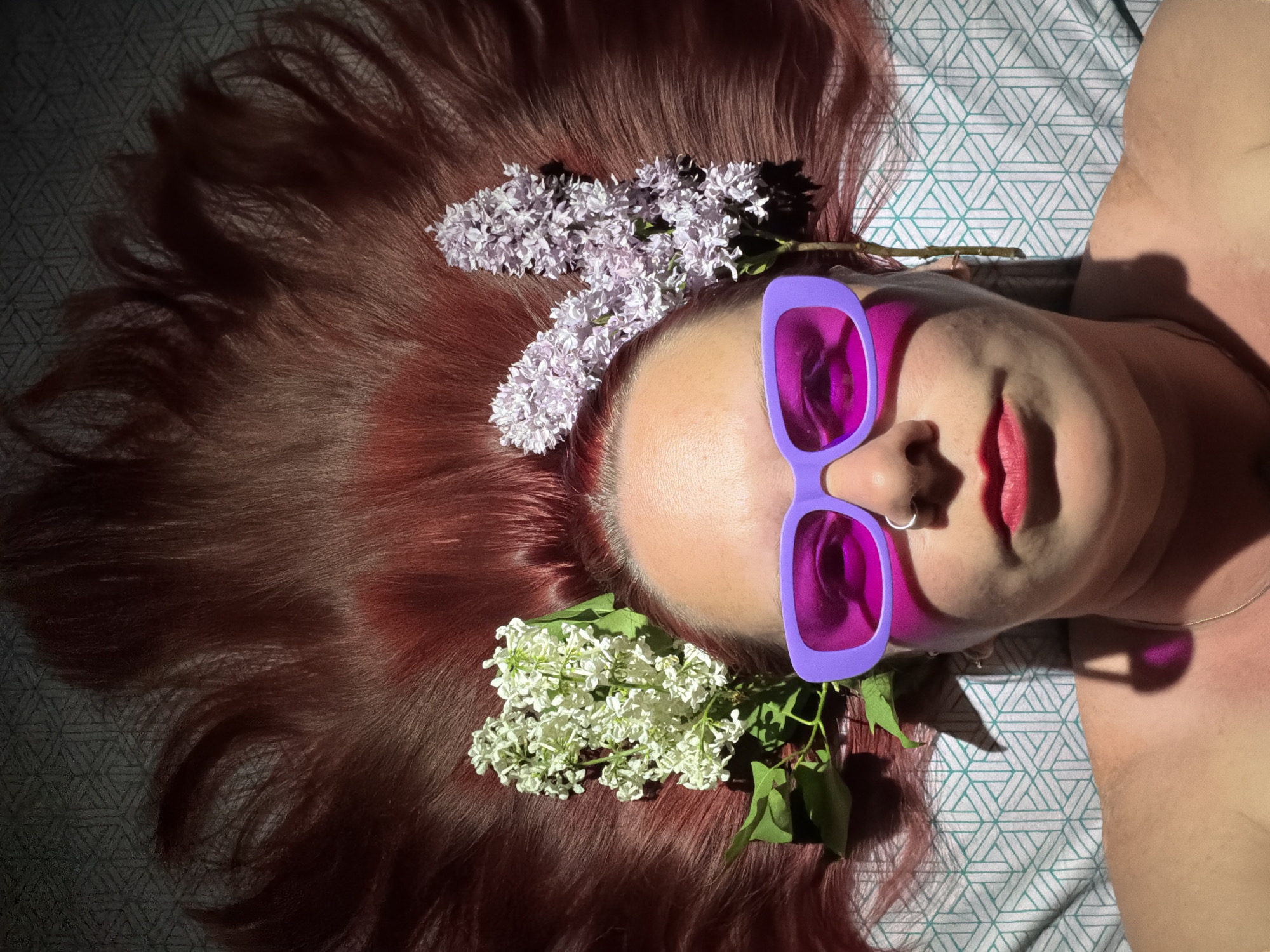 The image shows a person lying on a patterned surface, possibly a bedsheet with a triangular geometric pattern. The person has reddish-brown hair spread out around their head, with two clusters of white and purple lilacs placed in their hair. The person is wearing purple-framed sunglasses with magenta lenses. The person has fair skin, red lipstick, and a nose ring. The location is not specified and is unknown.