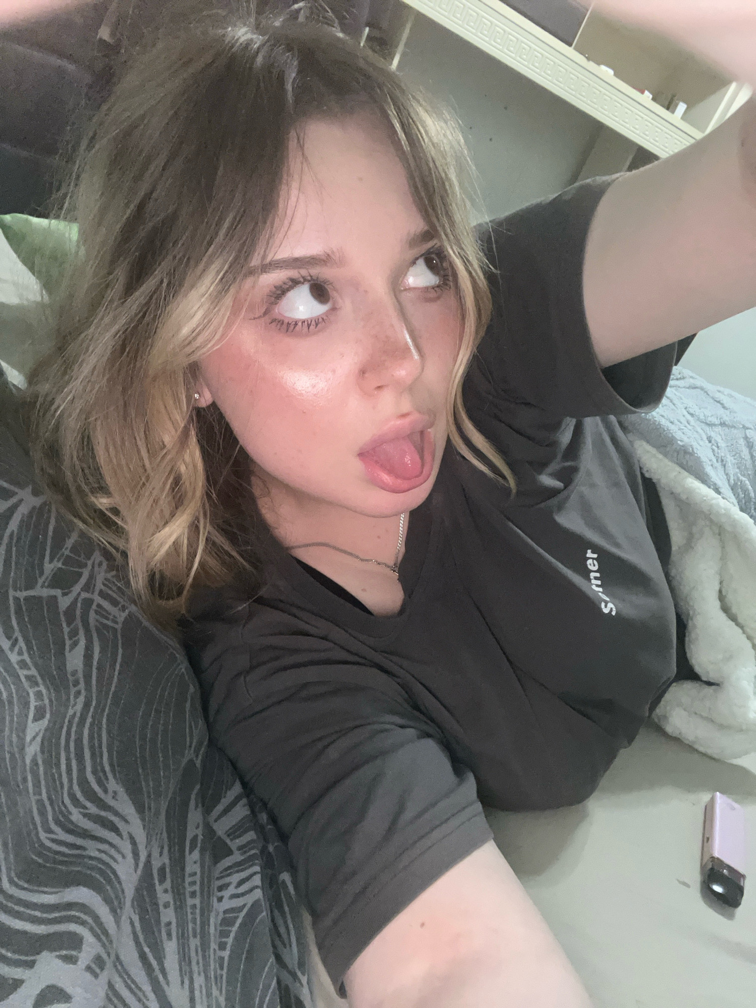 A young Caucasian woman with blonde hair and freckles is lying down and taking a selfie. She is wearing a dark grey t-shirt. Her tongue is sticking out and her eyes are looking upwards. She is lying on a bed or couch with a patterned pillow on her left and a fluffy white blanket on her right. A small pink electronic device is on the surface to her right. The background appears to be an indoor setting, possibly a bedroom, with shelves visible above. There is no indication of the city of Nizhny Novgorod, Russia, in the image.