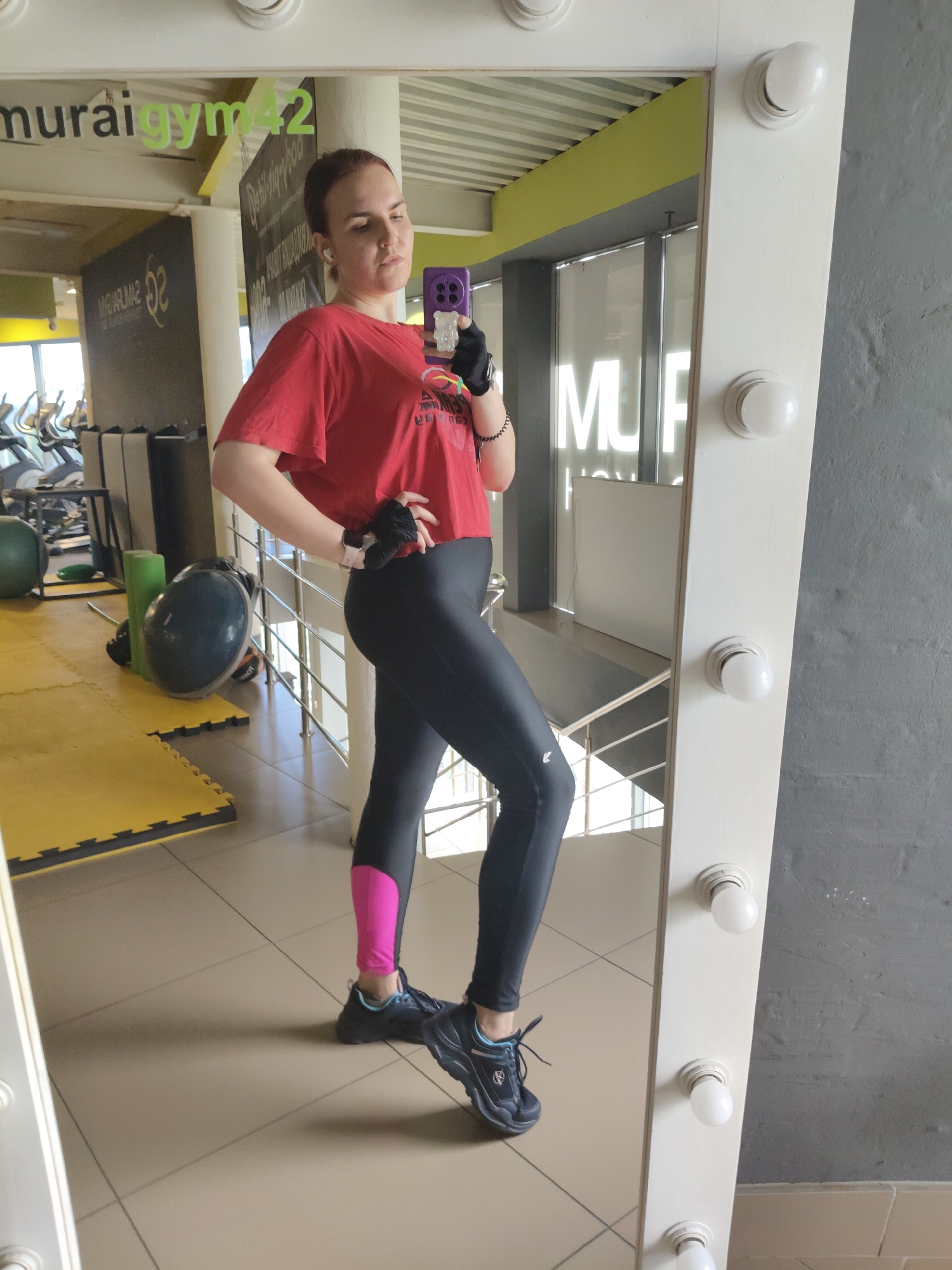 A woman is taking a mirror selfie in a gym. She is wearing a red cropped t-shirt, black leggings with a pink accent on the lower legs, and black athletic shoes. She has black gloves on and is holding a purple phone in her right hand. The gym appears to be well-lit, with exercise equipment visible in the background. The gym's name, "muraigym42," is visible on a wall in the reflection. The floor is tiled in a beige color. The overall environment suggests a typical fitness facility.