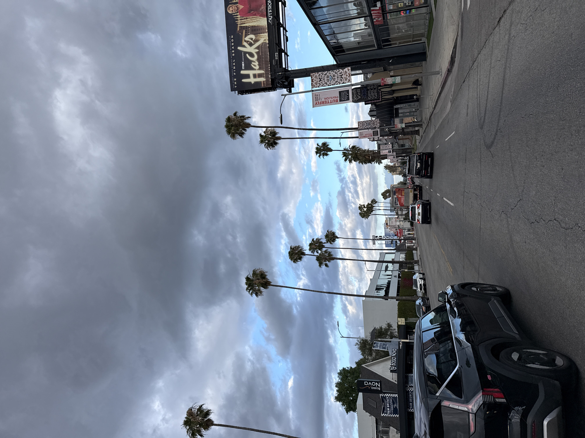 The image captures a street scene in Los Angeles, United States of America, with palm trees lining the road under a cloudy sky. A prominent billboard advertises the final season of "Hacks." Several vehicles are visible on the street, including a black SUV parked in the foreground. Signage for "Bossa Nova" and "Butterfly Pavilion" are also present.
