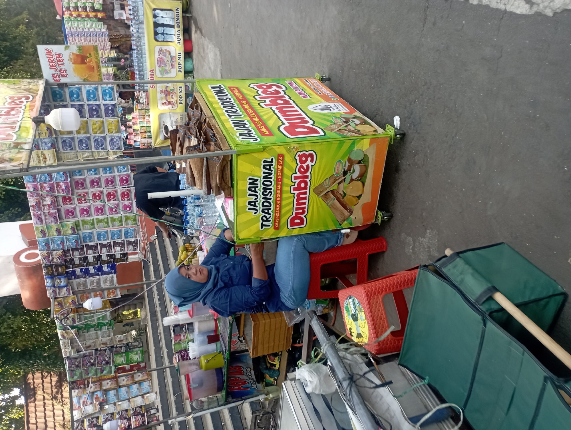The image depicts a street scene in Nganjuk, Indonesia, likely in a market or roadside area. A woman wearing a blue hijab and a blue denim shirt is seated on a red plastic stool in front of a bright green food cart. The food cart is decorated with images of what appear to be traditional Indonesian snacks, labeled "Jajan Tradisional" and "Dumbleg". The cart also has text indicating it is "Khas Nganjuk Gondang - Ngemplak", specifying its origin within Nganjuk. Other items visible include bottles of water, blenders, and various packets of food items displayed on shelves behind the cart. In the background, there are more stalls and structures, suggesting a bustling commercial environment. The ground is paved asphalt, and the lighting suggests it is daytime.