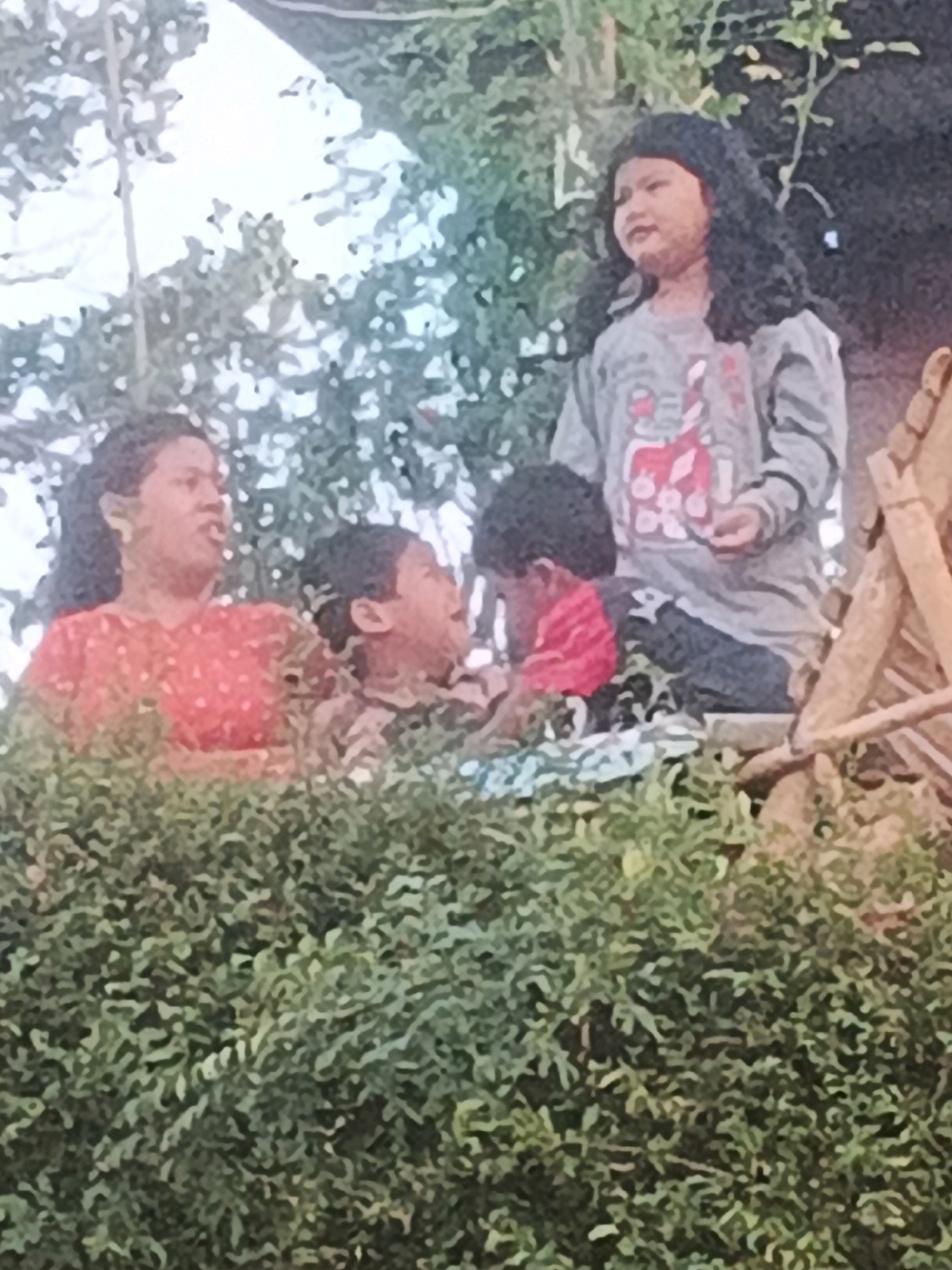 The image depicts a group of people, likely a family, outdoors in what appears to be a somewhat overgrown, natural setting. In the foreground, dense green foliage obscures the lower portion of the frame. Behind the foliage, a woman wearing a red, polka-dotted top is visible on the left, her mouth open as if speaking. To her right, a child with dark hair is looking upwards. Further to the right and slightly behind, another child with long, dark, curly hair is looking to the side, wearing a grey long-sleeved shirt with a graphic design. The background consists of trees and a light-colored sky, suggesting it is daytime. There are also some wooden structures or branches visible on the right side of the image. The image quality is somewhat grainy and lacks sharp detail, making precise identification of features difficult. There is no specific information in the image that places it in Keji, Indonesia.