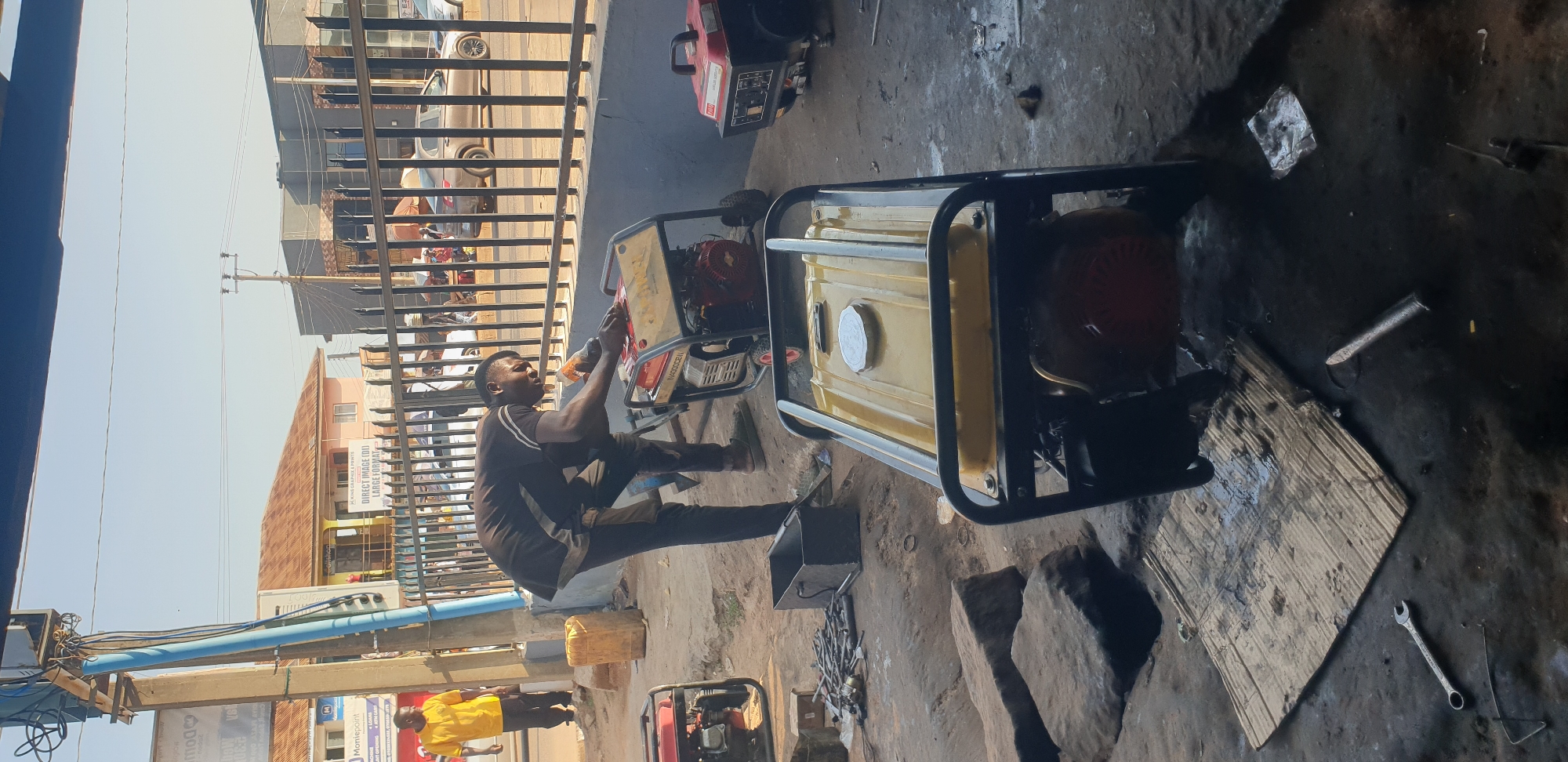 In Sabon Tasha, Nigeria, a mechanic is working on a portable generator in an open-air workshop. The scene is littered with tools, spare parts, and other generators. The workshop appears to be located on a dusty street with a building and shops in the background. A second individual in a yellow shirt stands nearby, observing. The sky is clear and the sun is shining, indicating it is daytime.