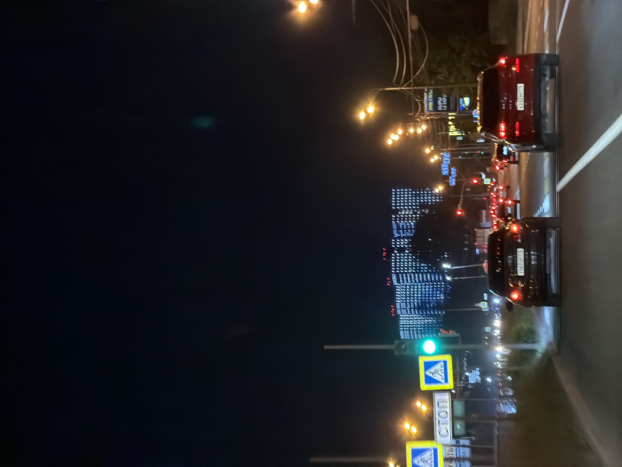 The image shows a nighttime view of a city street with traffic and a large illuminated building in the background. The perspective is from a vehicle, looking forward. Several cars are visible on the road, some with their headlights on. Traffic lights are also present, with a green light illuminated at one intersection. Road signs are visible, including a "STOP" sign and pedestrian crossing signs. Streetlights line the road, casting a warm glow. The large building in the background is covered in a pattern of blue and white lights, creating a distinctive facade. The overall scene depicts a busy urban environment at night.
