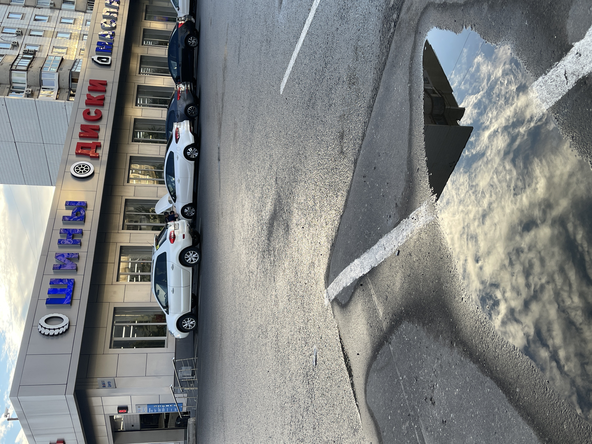 The image shows a street-level view of a commercial building with signage in Russian, likely in Moscow, Russia, as suggested by the Cyrillic alphabet. The building appears to be a tire and wheel shop, with the words "ШИНЫ" (Shini - Tires) and "ДИСКИ" (Diski - Wheels) prominently displayed on its facade. Several cars are parked in front of the building, including a white SUV and other sedans. The foreground of the image features a wet asphalt surface with a large puddle reflecting the sky, clouds, and parts of the surrounding buildings. A white line is painted on the asphalt, extending into the puddle. The sky is partly cloudy. The overall scene depicts a typical urban environment, possibly during or after rainfall.
