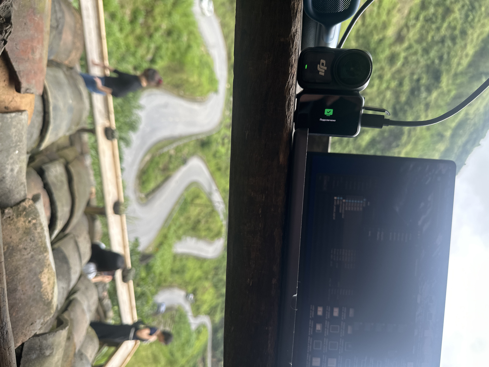 The image shows a laptop with a DJI action camera connected, positioned on what appears to be a wooden beam or ledge. The laptop screen displays program code or file directories. Through the background, a winding road through lush green terrain is visible, suggesting a mountainous or hilly location. On the left side, a roof made of tiles is seen, and several blurred figures can be distinguished walking on the roof with a winding road behind them. Based on the provided information, this scene is located in Yên Minh Commune, Vietnam.