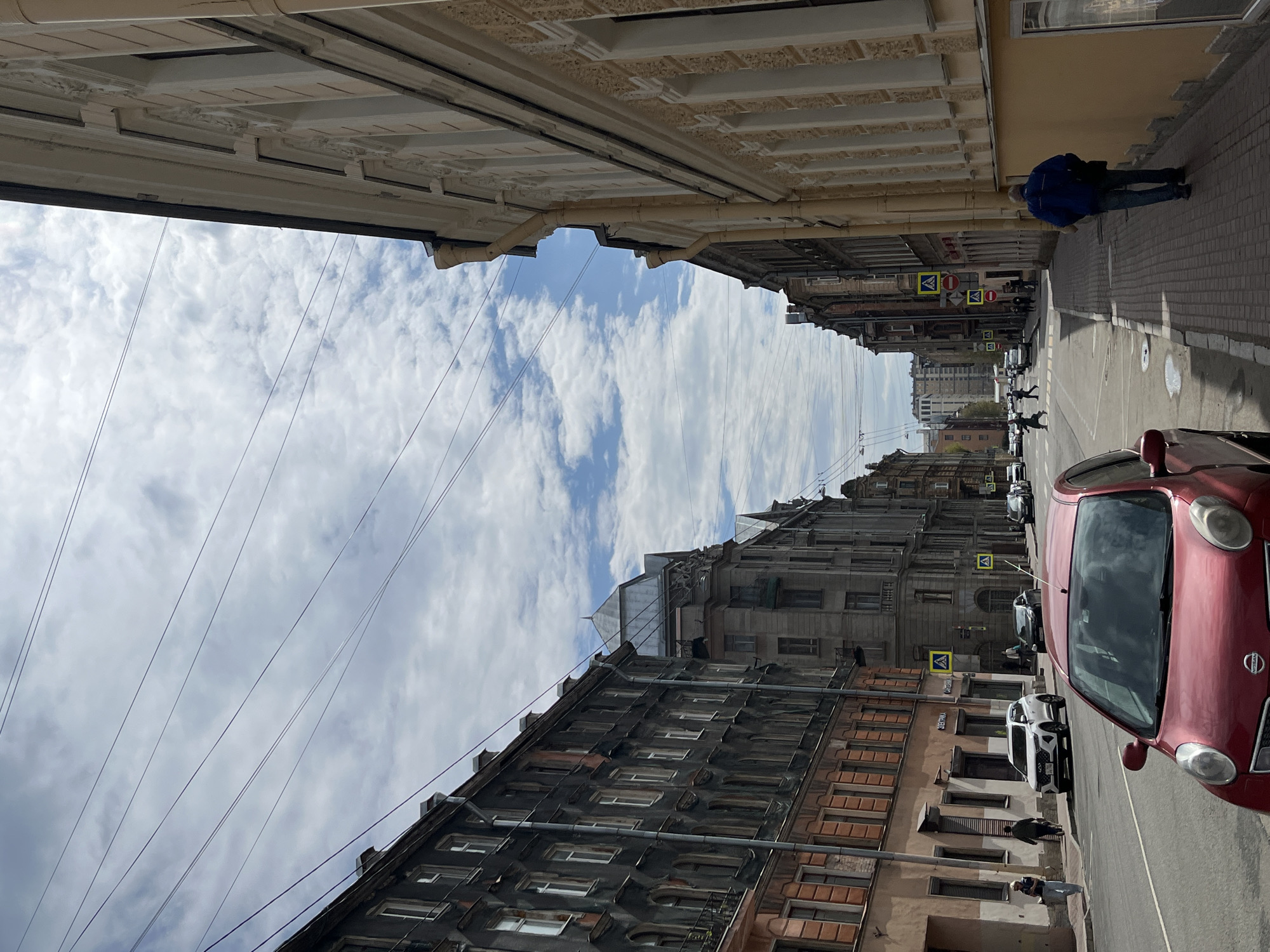 The image captures a street scene in Saint Petersburg, Russia. Buildings line both sides of the street, showcasing a mix of architectural styles and colors. Electrical wires stretch across the sky, framed by a cloudy sky above. A red car is parked on the right side of the street, partially obscuring the view further down the road where several other cars are parked. Pedestrian crossing signs are visible along the street. A person wearing a blue jacket is seen walking on the sidewalk to the right. Other pedestrians are also visible along the sidewalk.