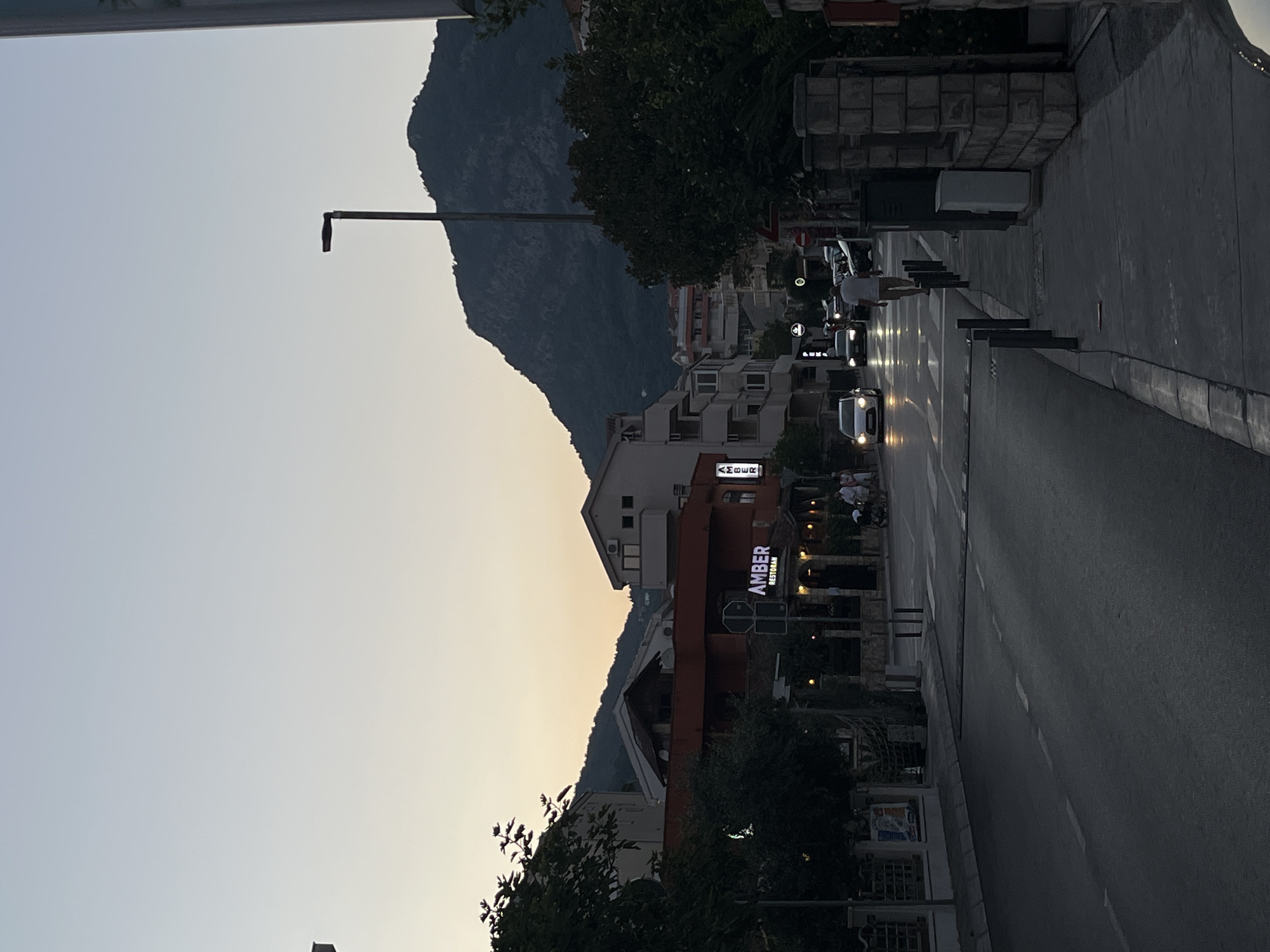 The image shows a street scene in Budva, Montenegro, at dusk. The sky is a pale gradient of blue and orange. On the left side of the street, buildings with red and white facades are visible, including a restaurant named "AMBER" with illuminated signage. Several cars are driving on the road, their headlights on. On the right side of the street, a stone wall and a sidewalk with lampposts and trash cans are present. A lone figure is walking on the sidewalk. In the background, a large, dark mountain looms over the city. The overall atmosphere is tranquil, with the streetlights beginning to illuminate the area.