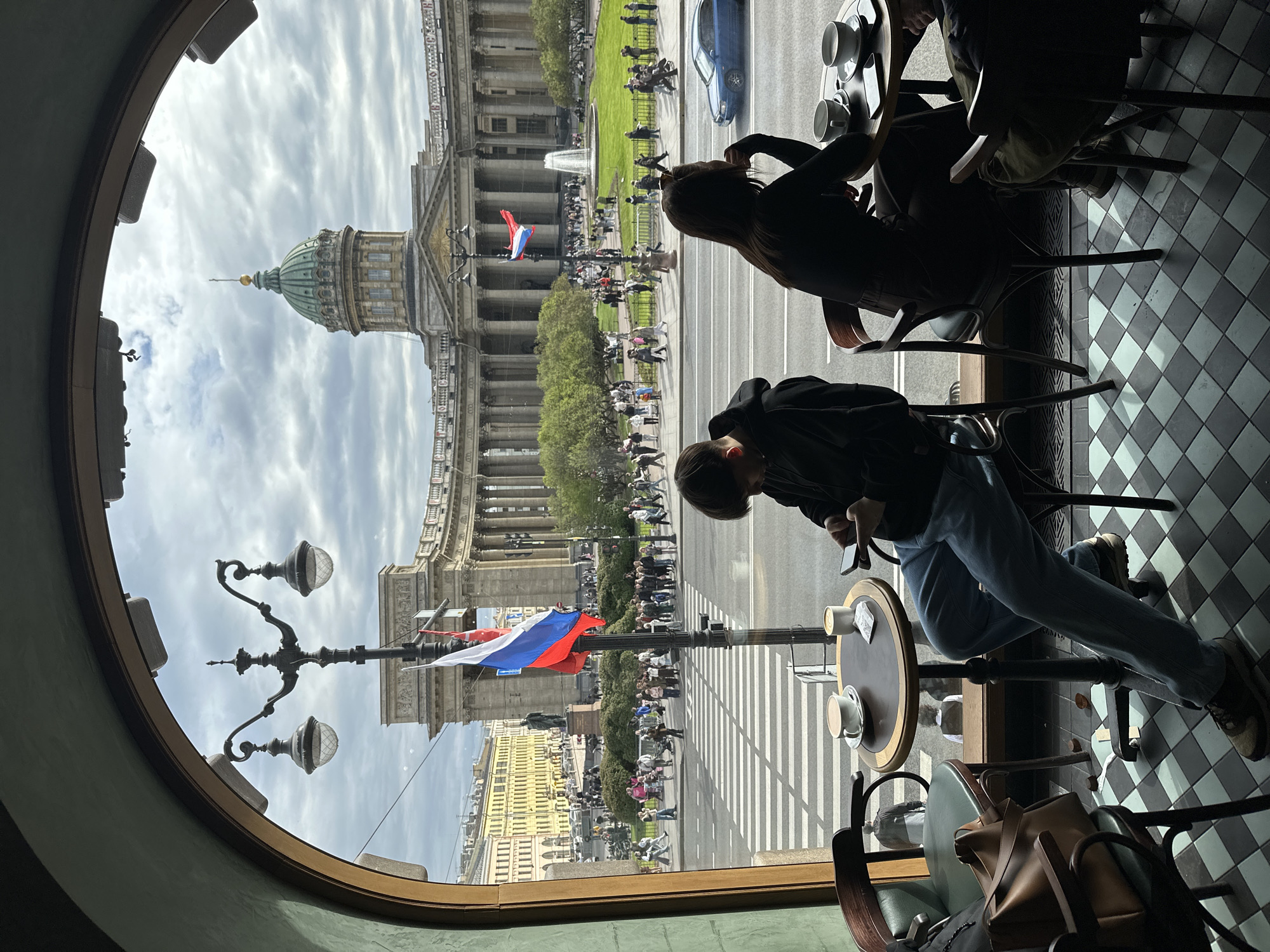 A cafe scene presents a view of Kazan Cathedral in Saint Petersburg, Russia, through an arched window. Inside the cafe, two individuals are seated at a table with coffee cups. One is focused on their phone. Outside, a pedestrian crosswalk stretches across a wide boulevard. The Russian flag is prominently displayed on a lamppost. A crowd of people is present, suggesting a public event or gathering. The weather appears overcast with cloudy skies. The floor of the cafe has a black-and-white checkered pattern. A backpack and chair are positioned near the foreground.