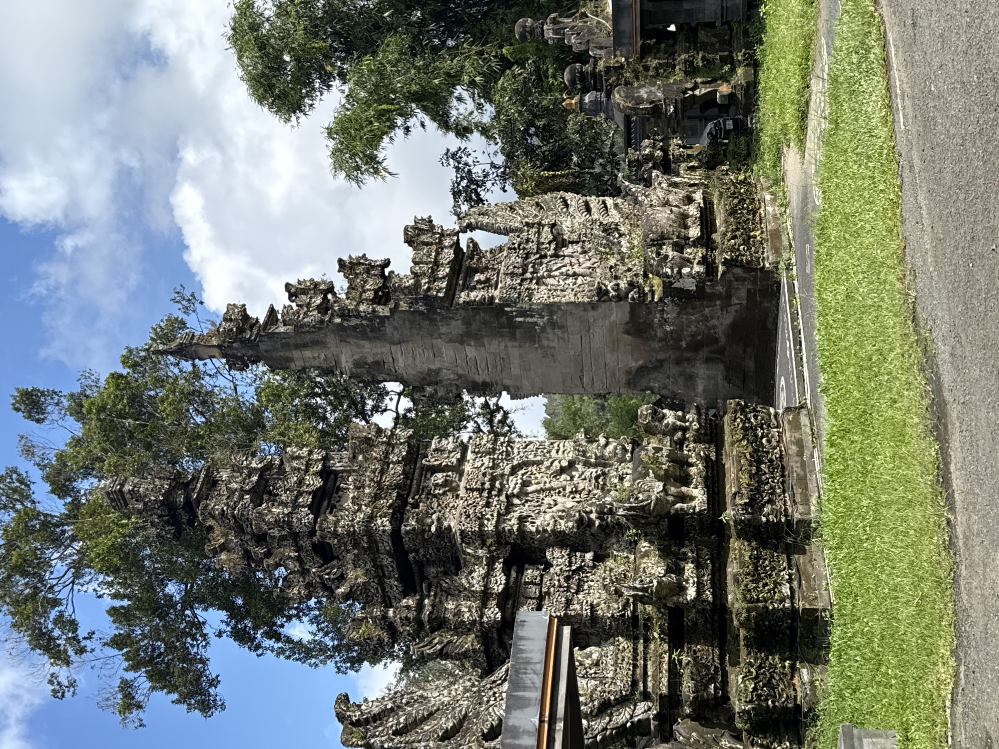 The image depicts the iconic Handara Gate, a Balinese Hindu gate located in Baturiti, Bali, Indonesia. The gate is a tall, grey, stone structure with intricate carvings and a passageway in the middle. Lush greenery, including trees, is visible both growing on the gate and in the surrounding environment. The sky is partly cloudy with a blue background. A grassy area and a paved road border the gate's base. The image is oriented sideways.