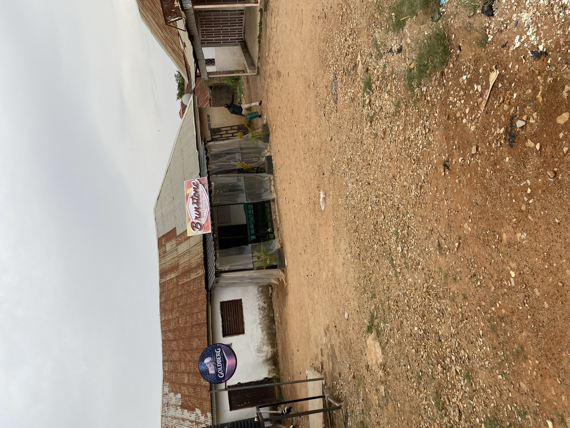 The image presents an outdoor scene in Barakin Akawo, Nigeria, under an overcast sky. The foreground and middle ground are dominated by an unpaved, reddish-brown dirt and gravel path, with scattered small rocks, some litter, and sparse green weeds.
On the left side of the path, a building with a corrugated metal roof, showing signs of rust, has white-painted walls and a dark, slatted window. In front of this building, a circular blue sign with white text reads "GOLDBERG LAGER BEER," mounted on a black metal stand.
Adjacent to this, a central building, likely a commercial establishment, also features a corrugated metal roof with significant rust and white walls. A prominent sign on its facade reads "Brunstone Wine & Smoker lounge" in orange, red, and white lettering. Below this, a smaller, partially visible sign appears to say "G&S SHOP." The front of the Brunstone lounge is covered with transparent plastic sheeting, and green potted plants are visible within.
In the middle distance, near the central building, a person is standing, clad in light-colored trousers and a dark top, holding a green bucket or container. Further into the background, another building with a metal gate is visible. The sky is a uniform light grey, indicating an overcast day. The overall setting suggests a local street or pathway in a semi-urban or rural environment.