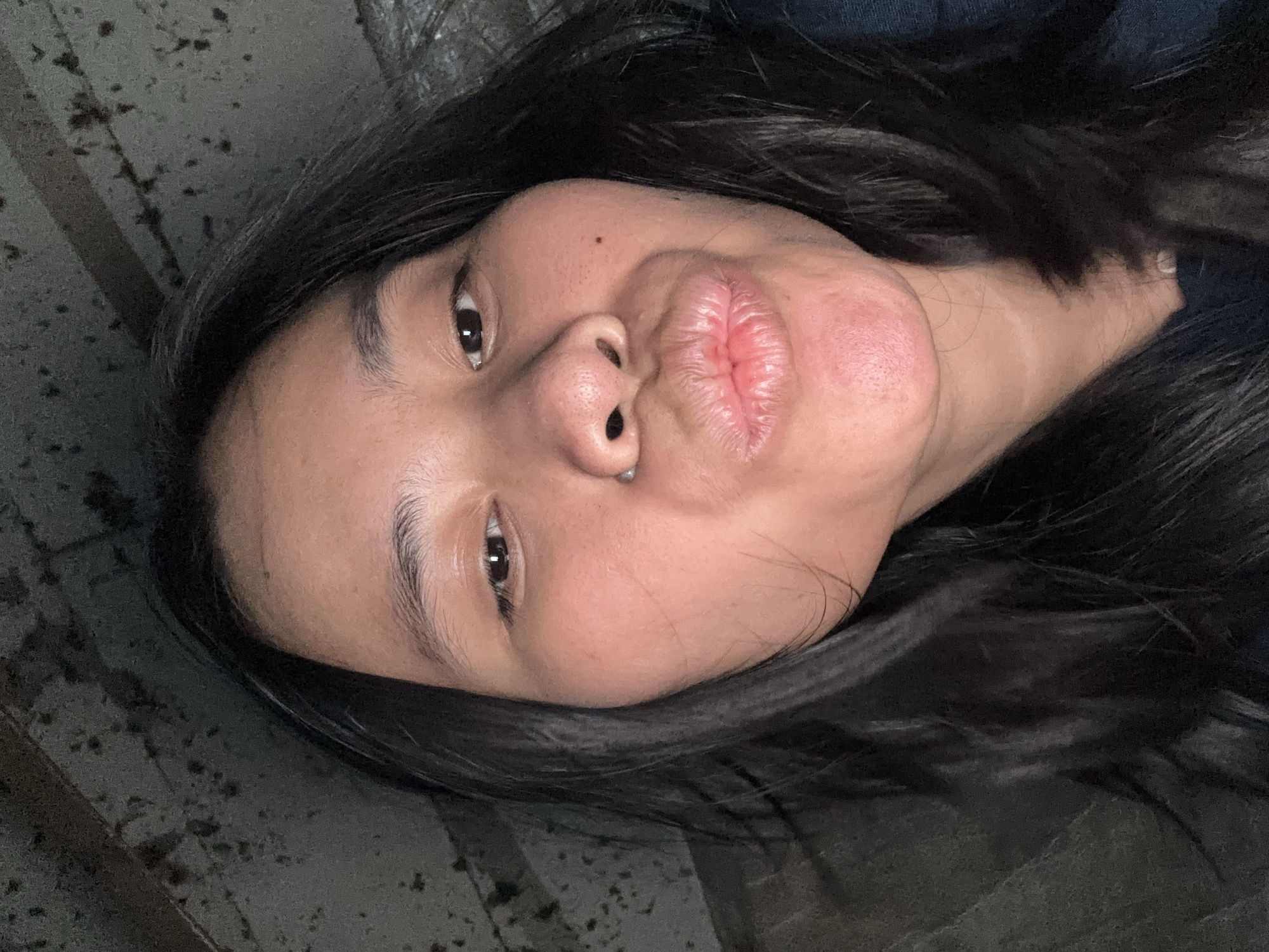 This is a close-up, horizontally oriented, low-angle, selfie of a young woman with long, dark hair. She is lying down, with her face angled upwards and towards the camera. Her lips are pursed in a kissing gesture. Her skin is light-skinned with some texture visible. She has dark eyes and dark eyebrows. Her hair is dark and falls around her face and shoulders.
The background is blurry and appears to be a dark, textured surface. There are some lighter, irregular spots or markings on this surface. The lighting suggests it may be indoors or at night, as there are no strong shadows indicating bright daylight.
The image appears to have been taken in East Jakarta, Indonesia, though no specific location cues are visible within the image itself. The overall mood conveyed is playful and casual. There is no visible text in the image.