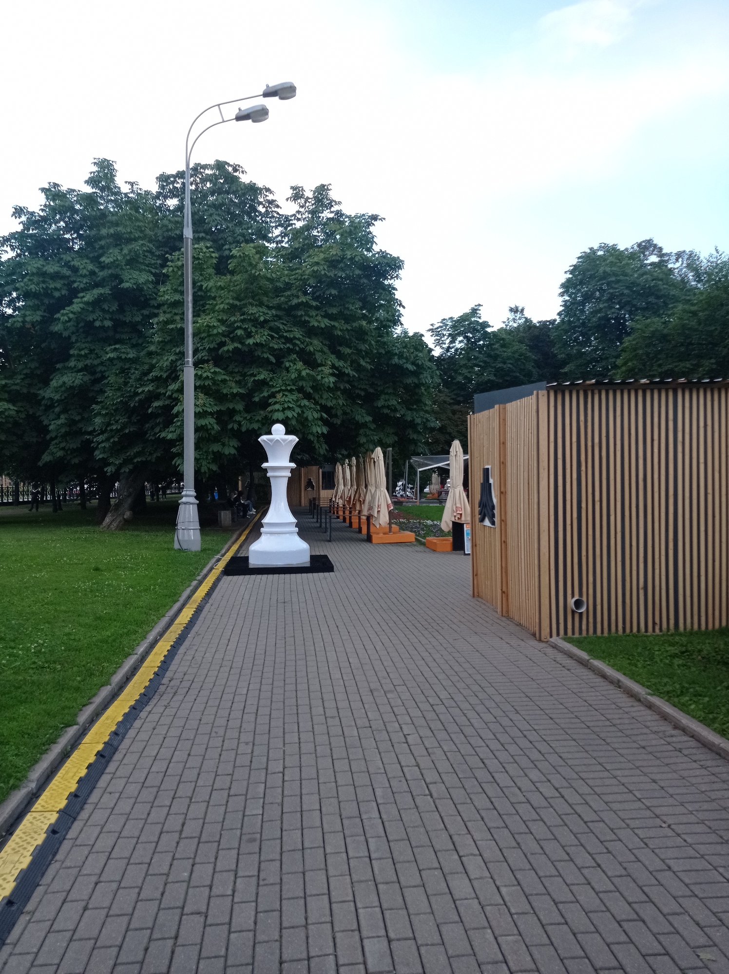 In the city of Moscow, Russia, a paved walkway in a park features a large white chess queen statue as its focal point. The statue stands on a black base next to the walkway, which is bordered by a grassy area on the left and a wooden structure with a ribbed facade on the right. A double-headed street lamp stands tall behind the chess piece. Rows of closed, beige-colored umbrellas are set up along the walkway, suggesting an outdoor seating area for a cafe or restaurant. In the background, lush green trees form a dense canopy, and the sky is a pale blue with a hint of clouds. A few individuals can be observed in the distance, walking on another path within the park. The overall scene depicts a public space with decorative elements and amenities for visitors.