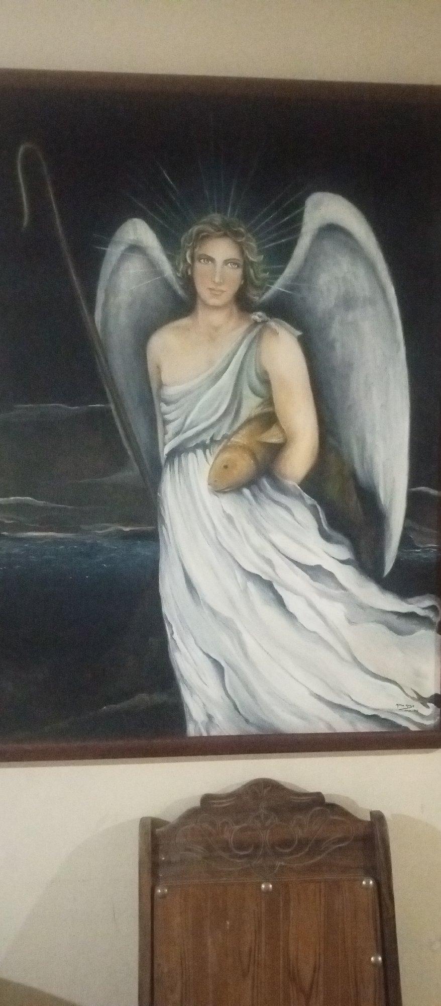 The image depicts a framed painting hanging on a wall. The painting shows a person with wings and a halo, wearing a white robe and holding a fish. They are positioned near water. A staff is visible behind the figure. Below the painting, a carved wooden chair is partially visible. The location is identified as the city of Tinaquillo, Venezuela.