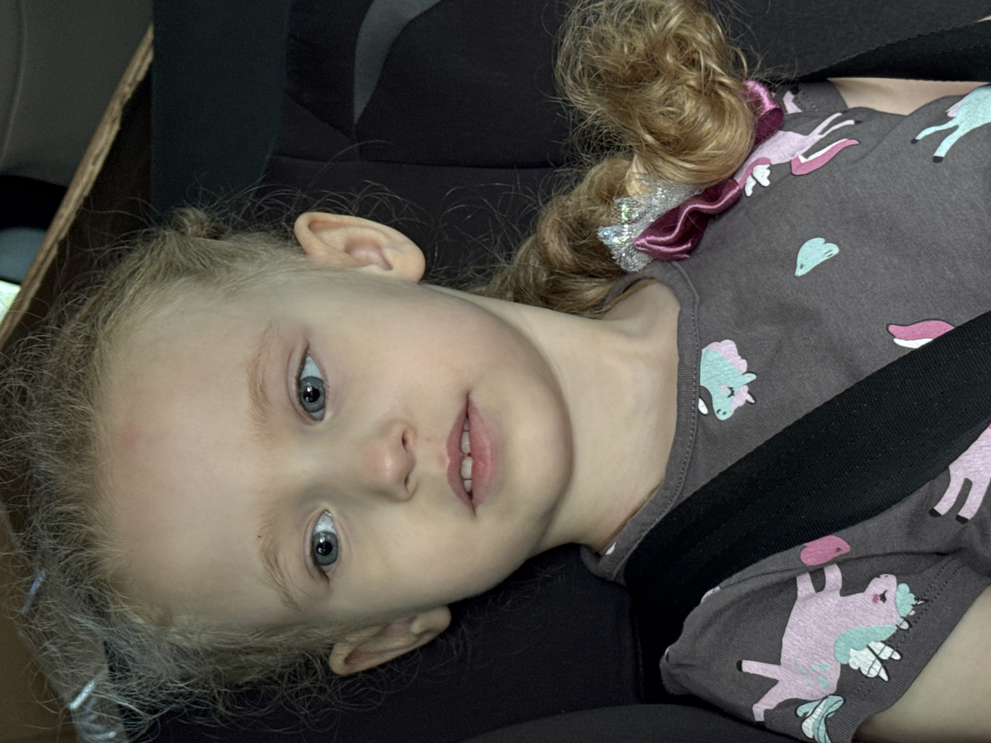 A close-up, slightly tilted portrait shows a young child, likely a girl, secured in a dark-colored car seat. The child has fair skin, prominent light blue or grey eyes, and curly blonde or light brown hair. Her expression is neutral, with her mouth slightly open. Her hair is adorned with a decorative pink and silver sequined ribbon on the right side of the frame. She is wearing a grey short-sleeved top patterned with various pastel unicorns and heart shapes in pink, light blue, and white. A black seatbelt stretches diagonally across her chest, indicating she is properly secured. The background reveals glimpses of a car's interior, including a dark-colored seat and what appears to be a light-brown wooden-like trim on the left. The lighting suggests it is daytime. The child is looking directly at the camera, implying a photograph is being taken while traveling by car. No text is visible in the image. This scene potentially takes place in Chaikovsky, Russia.