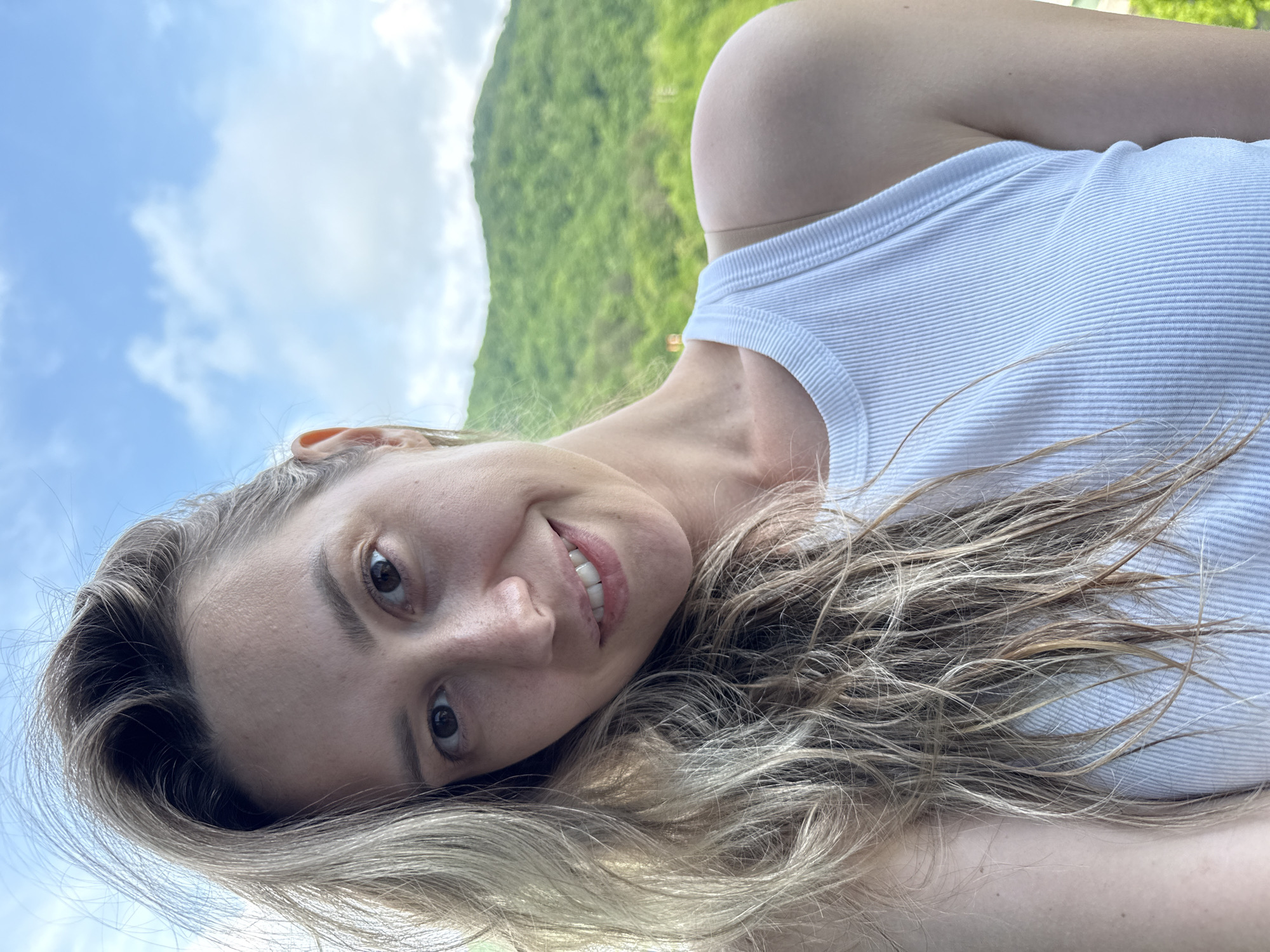 A medium shot selfie of a young Caucasian woman with blonde hair framing her face and shoulder. She is wearing a white ribbed tank top and smiling. Her eyes are brown and she has fair skin. The background consists of a green, tree-covered mountain slope under a cloudy blue sky, characteristic of the natural landscape near Nebug, Russia. The lighting suggests it is daytime.