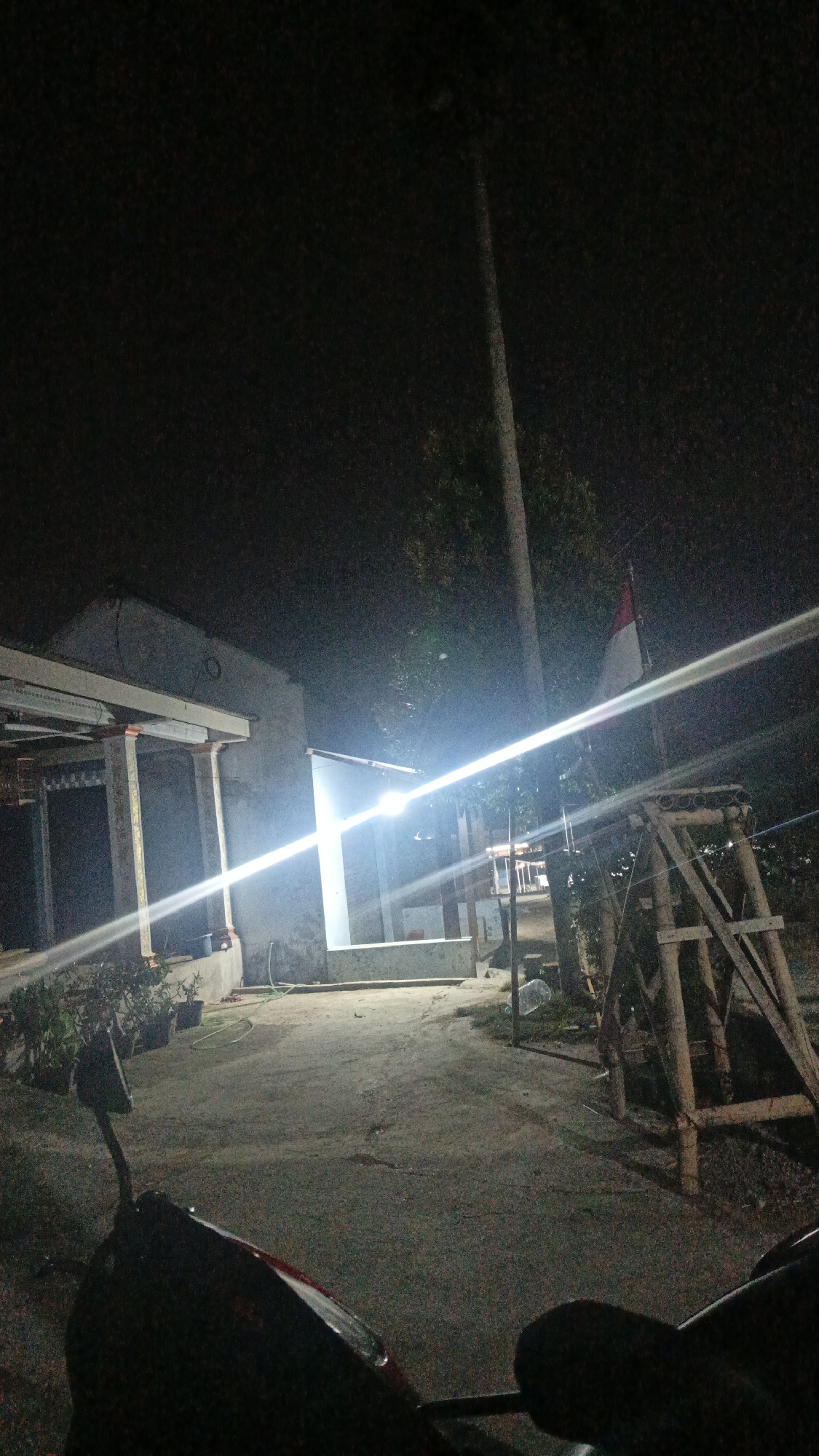 The image depicts an outdoor scene at night, likely in Sambi, Indonesia. A bright light source, possibly a streetlamp or powerful outdoor lighting, illuminates the central area, creating strong glare that stretches across the frame. To the left, a building with a covered porch supported by columns is visible. Potted plants are arranged along the front of this building.
In the middle ground, a white structure with a concrete base is illuminated by the bright light. A green hose lies on the ground in front of it. To the right of this structure, a flagpole stands with the Indonesian flag (red and white) flying. Adjacent to the flagpole is a wooden scaffolding-like structure.
The ground appears to be a paved pathway or road. In the foreground, the handlebars and part of the front of a dark-colored motorcycle are visible, suggesting the image was taken from the perspective of someone riding or parked on the motorcycle. The overall environment appears to be a residential or semi-rural area. The scene is dark, with the primary illumination coming from the artificial light source.