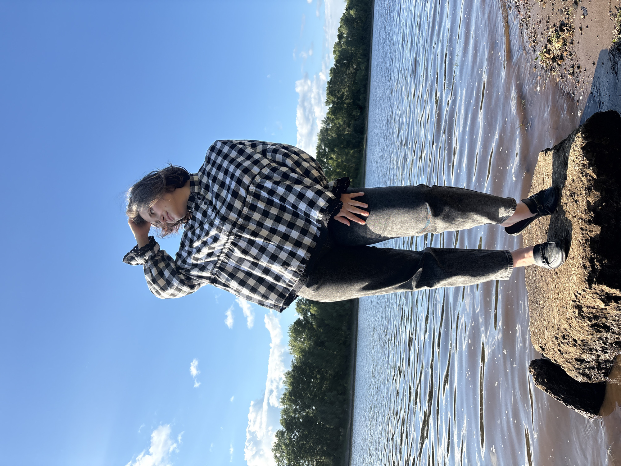 A person stands on a large rock at the edge of a body of water. The person is facing to the right, with their left hand on their head and their head tilted slightly. They are wearing a black and white checkered long-sleeved shirt and dark pants, with black loafers. The water is calm with ripples and reflects the bright blue sky with scattered white clouds. A line of green trees is visible across the water in the background. The ground around the rock is a mix of sand and small stones.