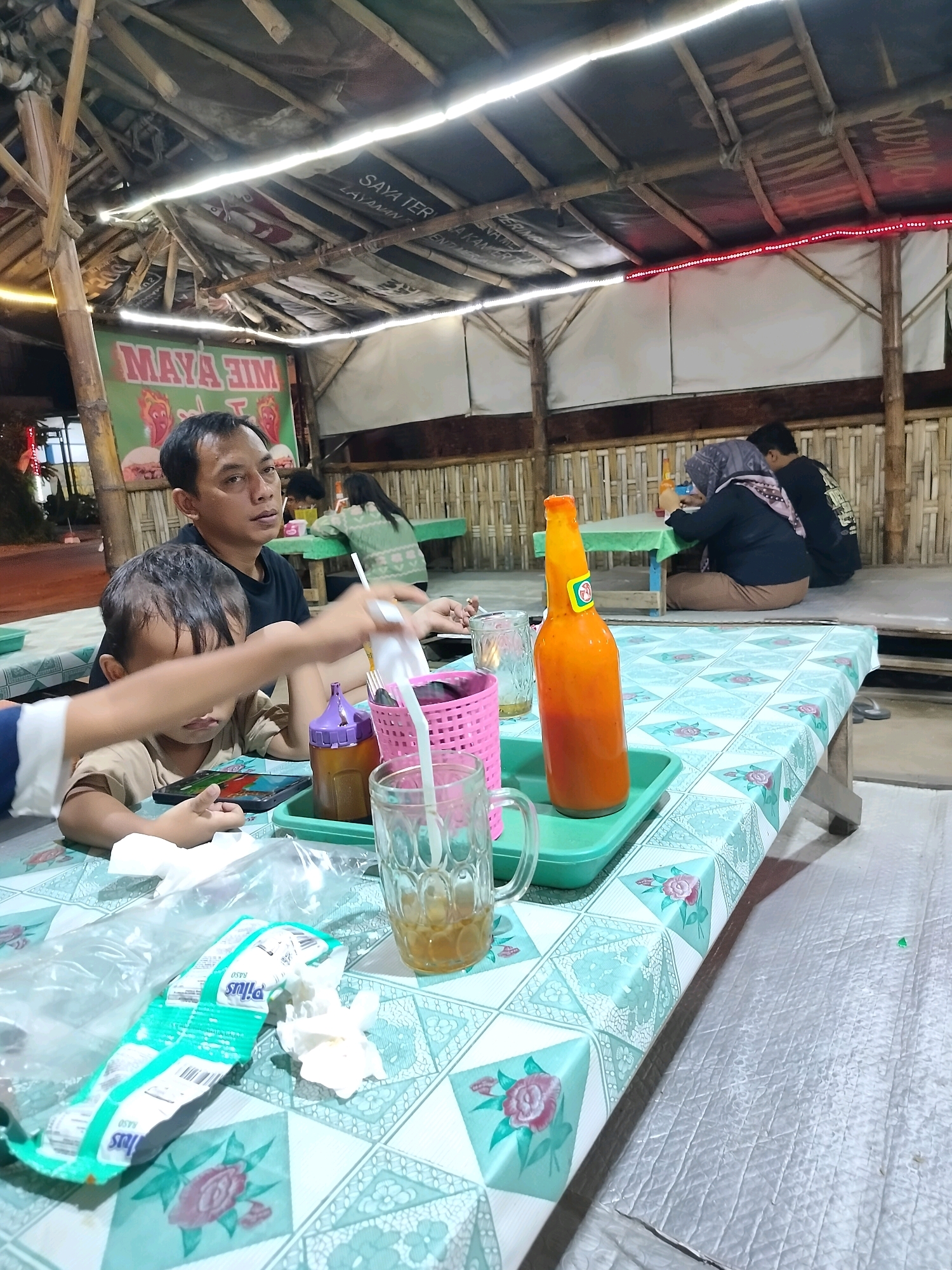 In Trowulan, Indonesia, a family is dining at a casual outdoor restaurant with a bamboo and thatch roof. A father, wearing a black t-shirt, sits with his young son who is looking at a mobile phone. The boy is reaching for something on the table. On the table, there are drinks in glasses, a bottle of orange sauce, and some crumpled napkins. In the background, other patrons are seated at tables. The environment appears to be a rustic, open-air dining establishment.