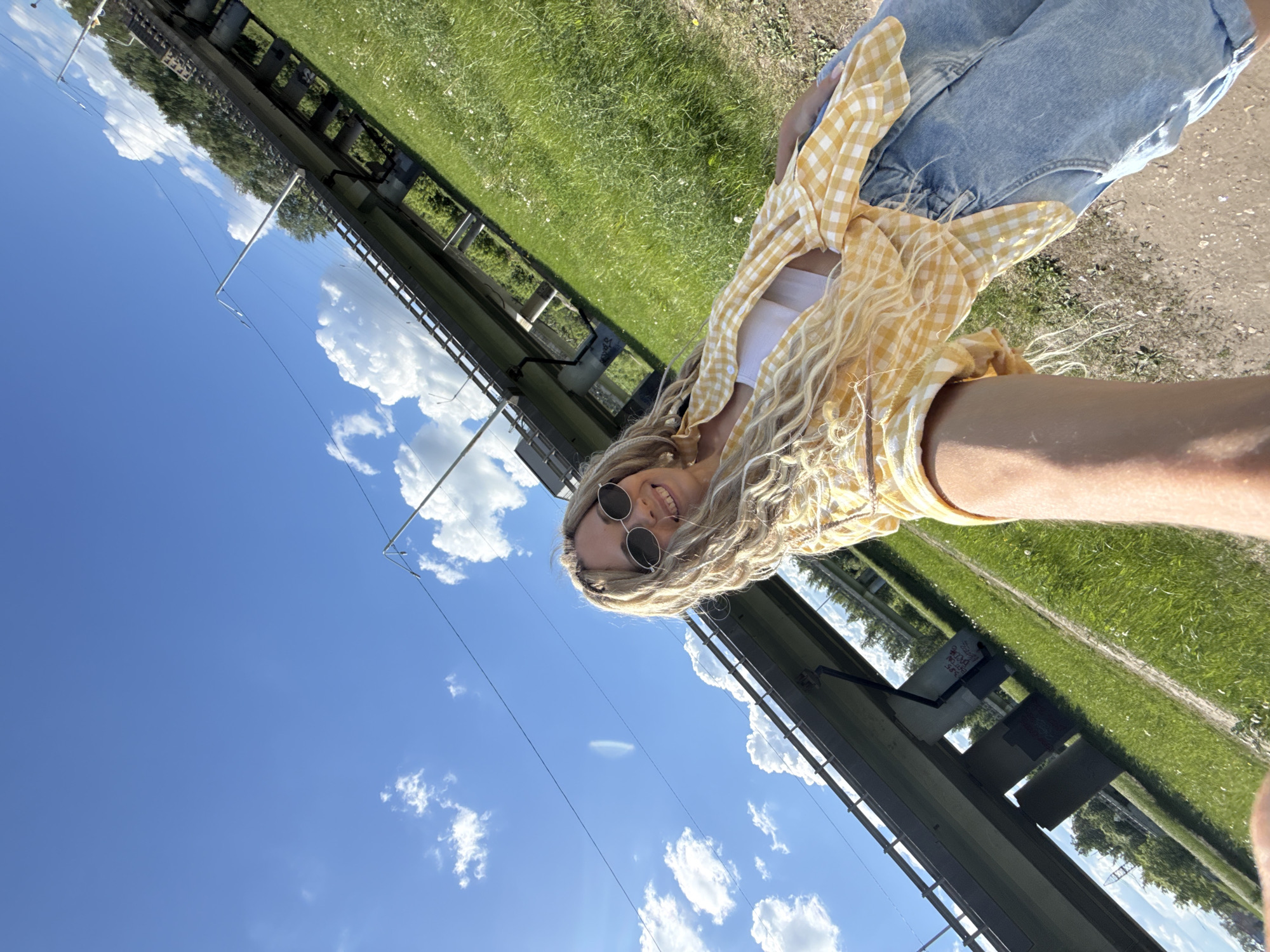 A young woman with blonde curly hair and wearing sunglasses is posing for a selfie. She is wearing a white top, a yellow checkered shirt, and denim jeans. The photo is taken from a low angle, capturing the clear blue sky with scattered white clouds. In the background, there is a metal structure and greenery, possibly indicating a bridge or elevated platform. The location is Заволжье, Russia, as indicated in the prompt.