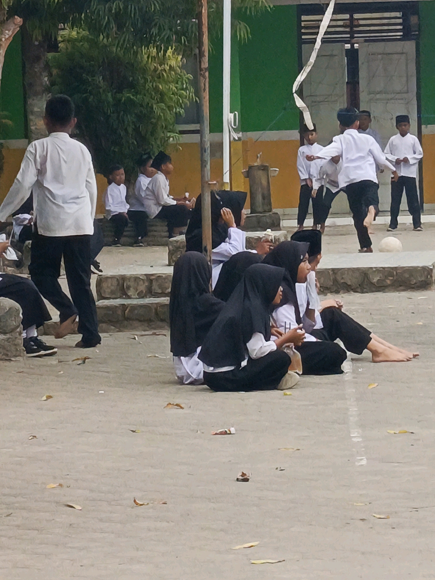 In an outdoor setting, likely a schoolyard in Megang Sakti II, Indonesia, a group of children and young people are gathered. Several individuals are seated on a paved area, some wearing black hijabs and white shirts, suggesting a school uniform. Others are standing or in motion.
In the background, a building with green and yellow walls is visible, with a wooden pole and a white banner or ribbon hanging from it. Some of the standing individuals are wearing white shirts and black pants, also appearing to be in uniform. One person in the background is in mid-motion, possibly participating in a game or activity, with a white ribbon trailing behind them. A white ball is visible on the ground near the standing figures. The ground is paved, with scattered leaves and debris. The scene suggests a daytime event, possibly a school activity or a cultural celebration.