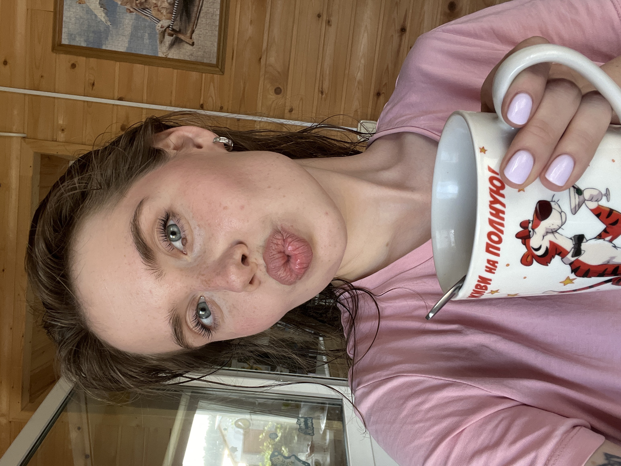 The image shows a young woman with wet brown hair and blue eyes, wearing a pink t-shirt. She is holding a white mug with a cartoon tiger and Russian text. Her lips are pursed in a kissing gesture. The background appears to be a wooden interior with a window that offers a view of houses and greenery, possibly a residential area in Chaikovsky, Russia. A framed picture is visible on the wooden wall. The overall scene suggests a casual, indoor setting.