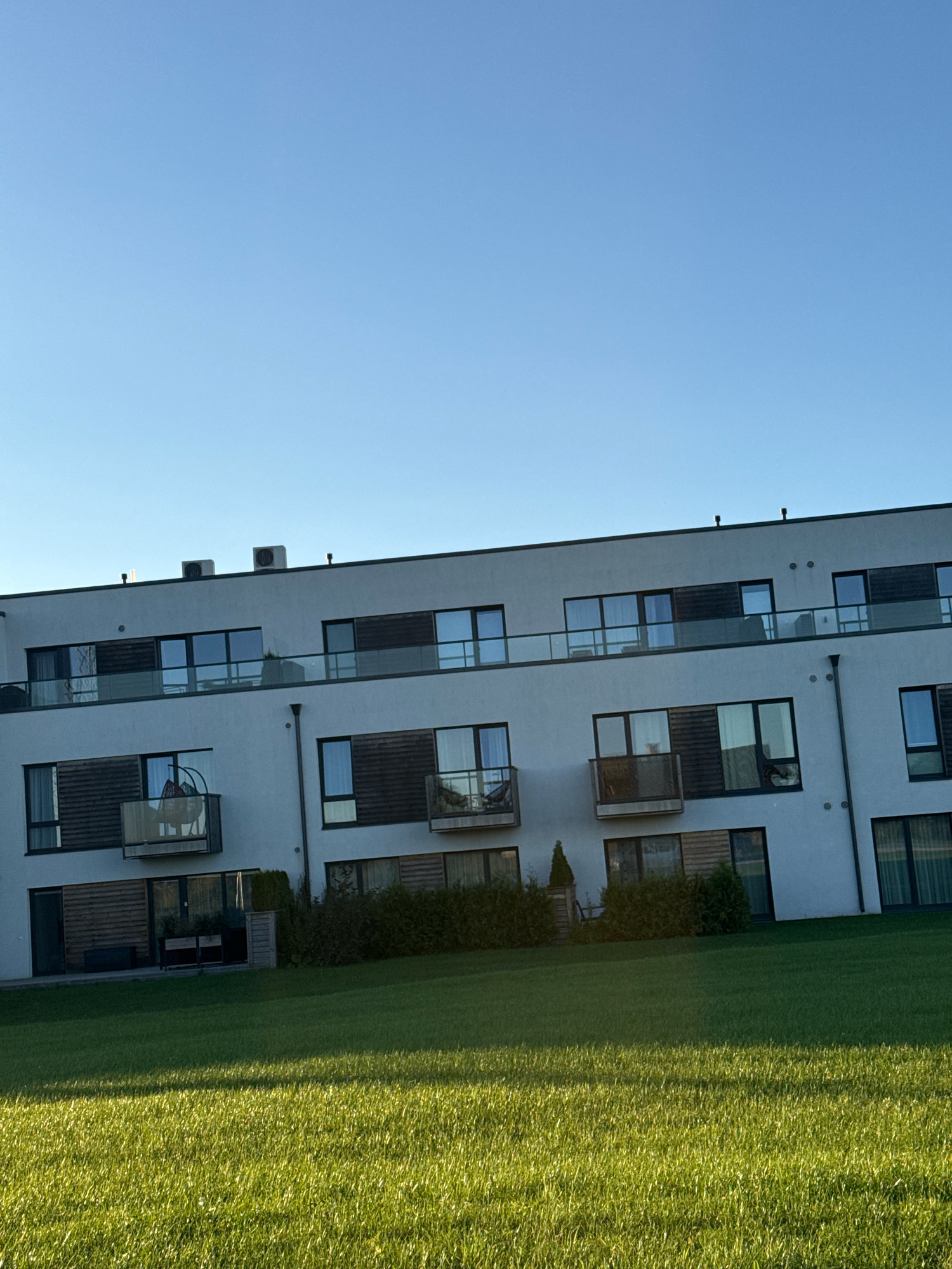 The image shows the exterior of a modern apartment building with a light-colored facade, large windows, and balconies. The building has several stories, and its architectural style suggests a contemporary design. Two outdoor air conditioning units are visible on the roof. In front of the building, there is a well-maintained green lawn, illuminated by sunlight, suggesting it is either morning or late afternoon. The sky is clear and blue, indicating fair weather. The building is situated in Järveküla, Estonia, as indicated by the metadata. There are no people visible in the image, and no specific actions or interactions are taking place. The scene depicts a serene and orderly residential environment.