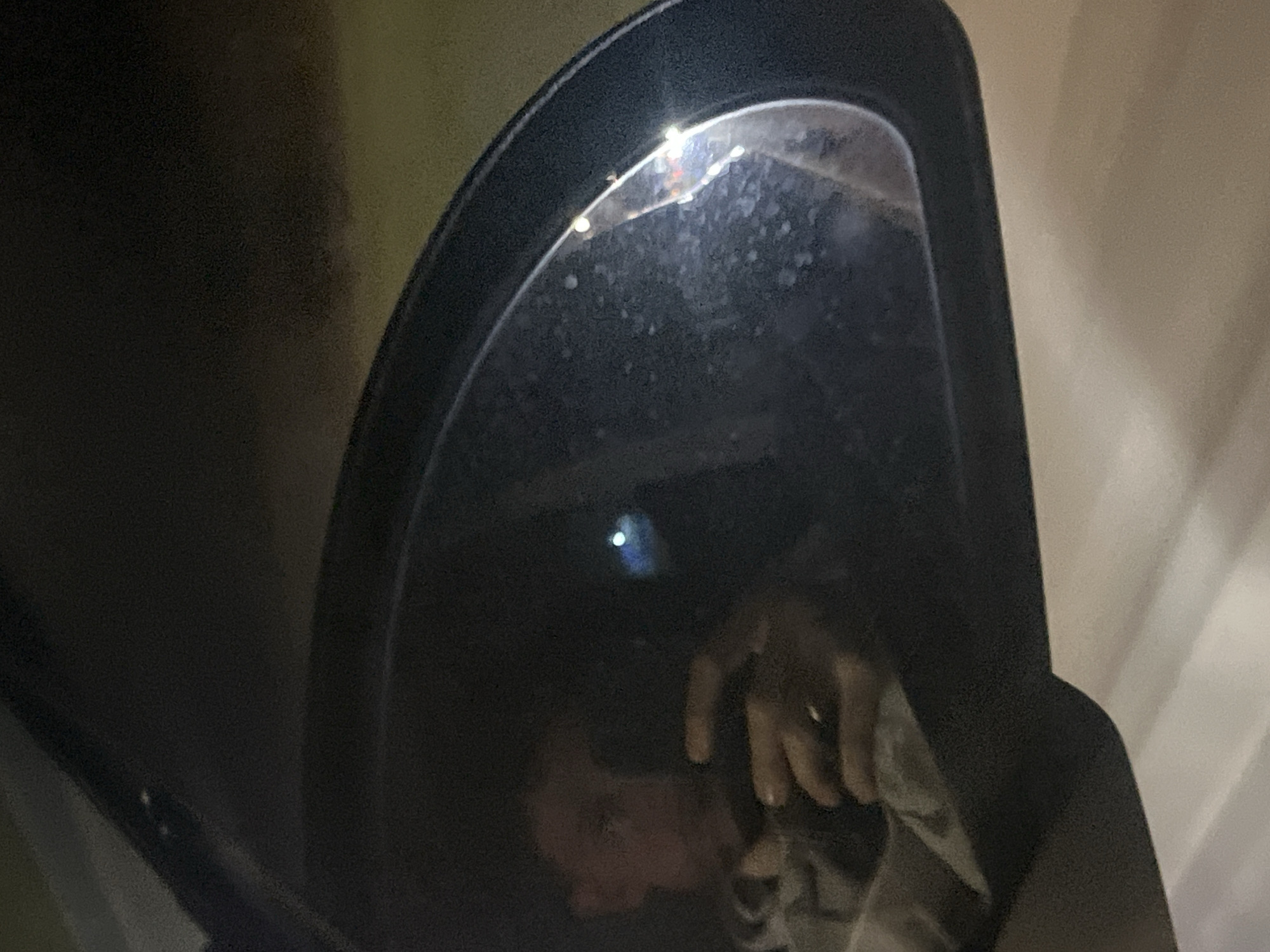 The image shows a partial reflection in a car side mirror. The reflection includes a person's face, a hand wearing a ring, and a portion of clothing. The background outside the car is blurry and indistinct, showing only vague shapes and light colors. The exact location in Sofia, Bulgaria cannot be determined from this image.