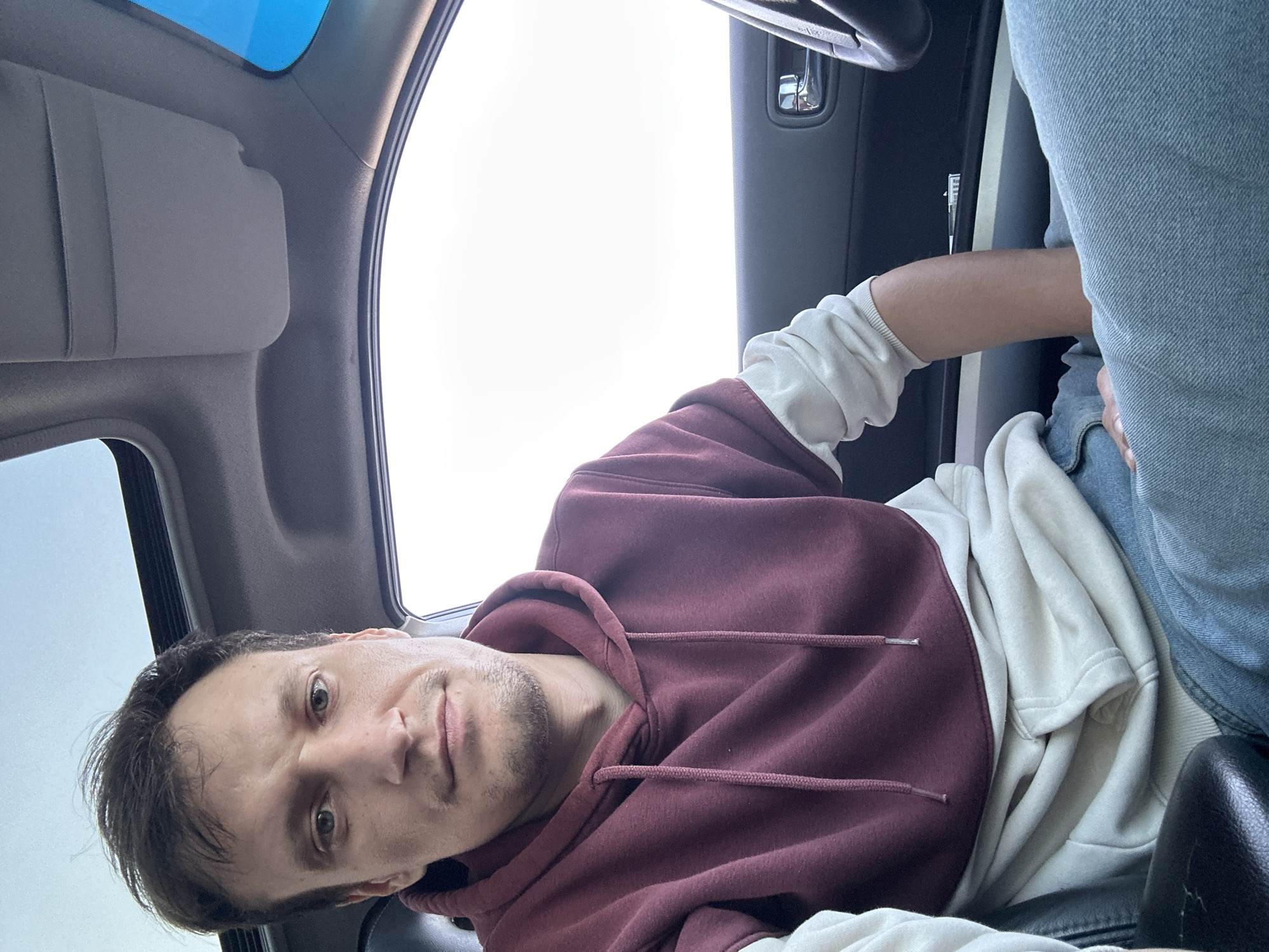 A man is sitting in the driver's seat of a car, looking towards the camera. He is wearing a burgundy and cream hooded sweatshirt and blue jeans. The car's sunroof is open, and the sky is visible through it. The interior of the car is visible, including the dashboard, steering wheel, and door panel. There is no indication of the specific location within the city of Kamianets-Podilskyi, Ukraine.